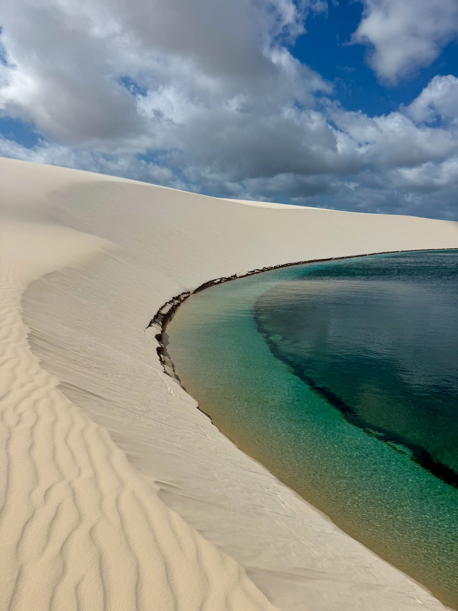 Rainwater pooled in silk — the impossible blue of a dune lagoon. A crystal-clear turquoise lagoon curves along the base of a sweeping white sand dune under a dramatic cloudy sky in Lençóis Maranhenses Santo Amaro