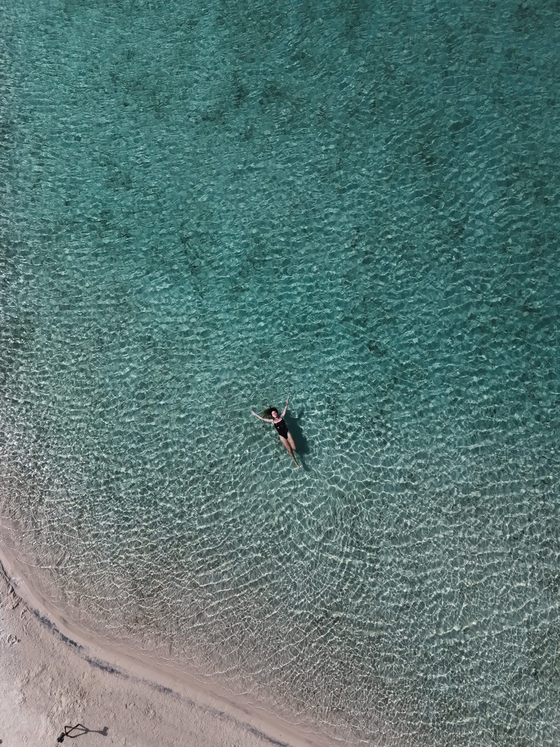 Aerial drone view of a woman in a black swimsuit floating in the crystal-clear turquoise shallows of Lençóis Maranhenses tours swimming lagoon, with white sand visible at the water's edge