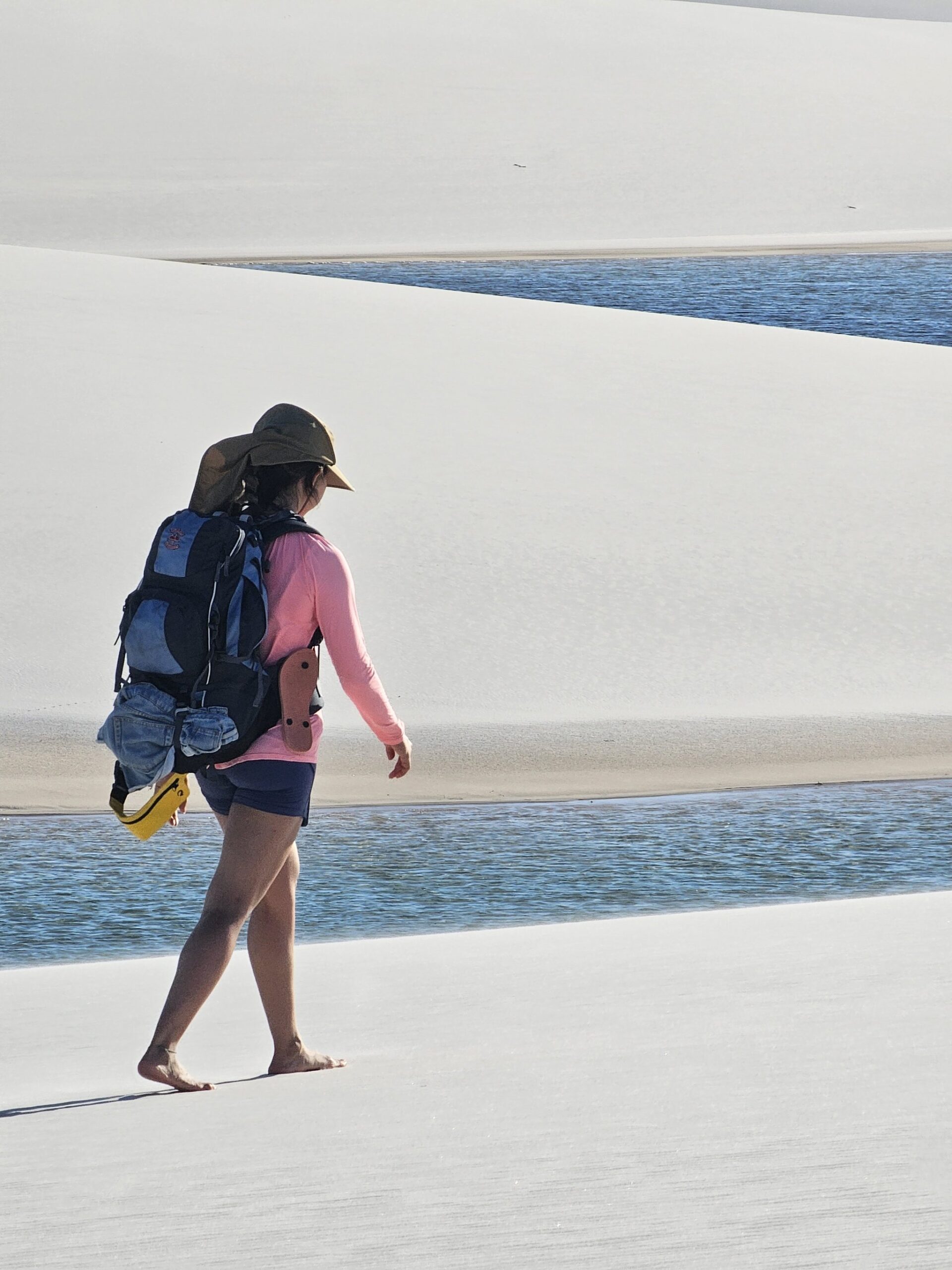 A backpacker walks barefoot across white sand dunes beside a crystal-clear rainwater lagoon during a Lençóis Maranhenses tour