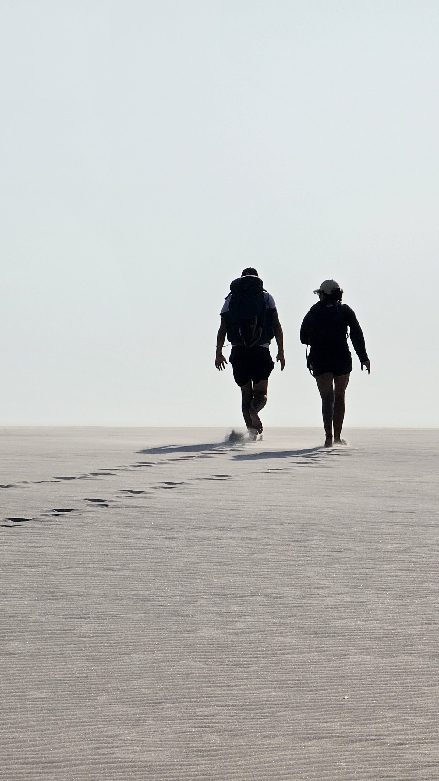 Two hikers with backpacks walking across vast white sand dunes on a Lençóis Maranhenses tour, leaving a trail of footprints behind them