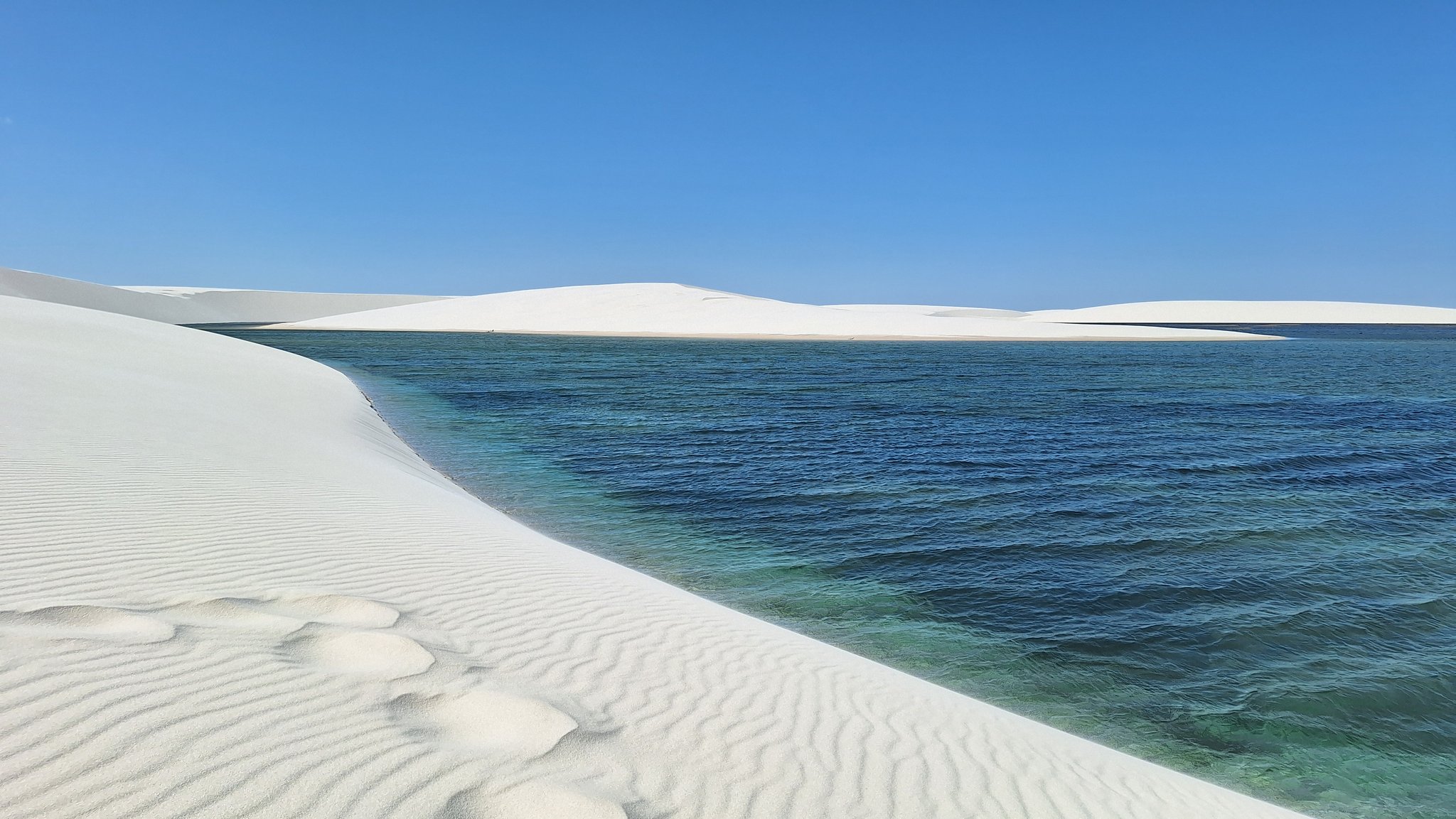 Rippled white sand dunes meeting a deep turquoise lagoon under a clear blue sky in Lençóis Maranhenses, Brazil