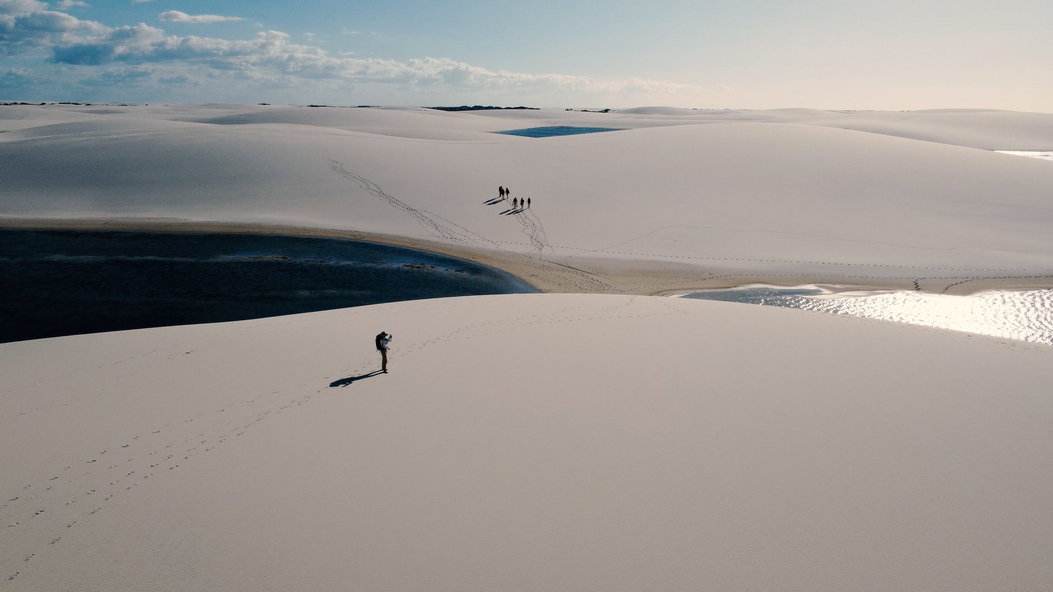 Aerial view of a lone hiker photographing the white sand dunes of a lençóis maranhenses trek, with dark freshwater lagoons curving across the landscape and a small group of trekkers visible in the distance