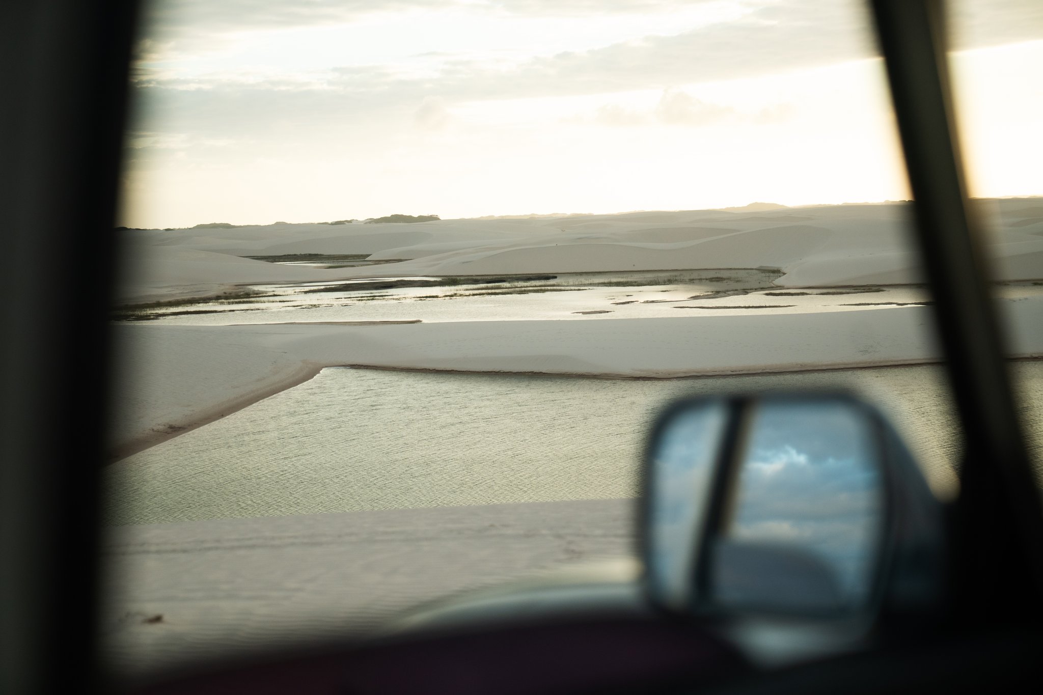Through the window: the dunes and lagoons appear, immense and unhurried. View through a 4x4 vehicle window of vast white sand dunes and shallow shimmering lagoons stretching to the horizon on the Lencois Maranhenses Trek from Atins, with a side mirror reflecting the sky