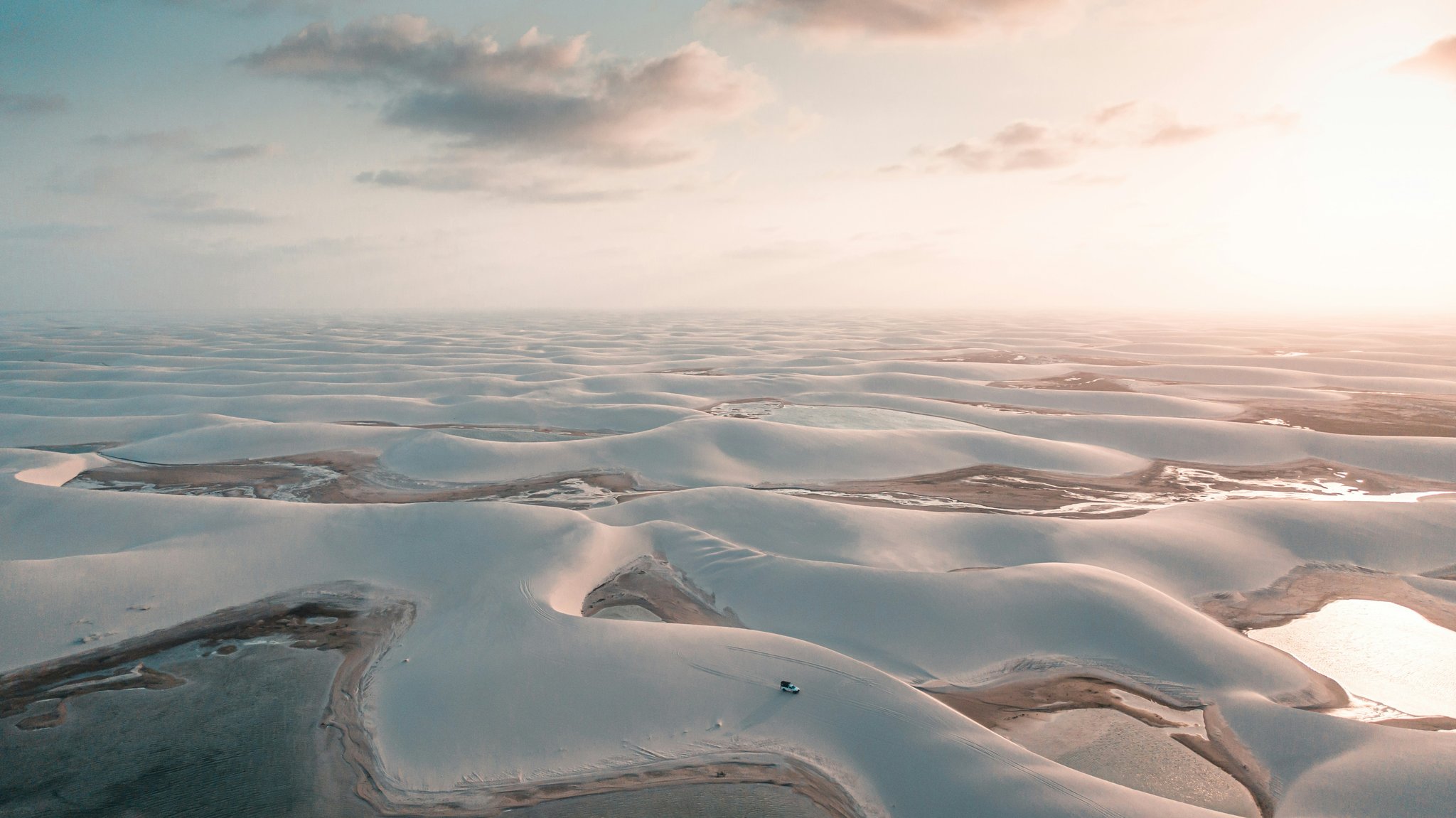 Endless dunes, scattered lagoons — and one small truck making its way through. Aerial view of the Lençóis Maranhenses Trek from Atins showing a tiny 4x4 vehicle crossing vast white sand dunes dotted with shallow freshwater lagoons under a warm glowing sunrise sky