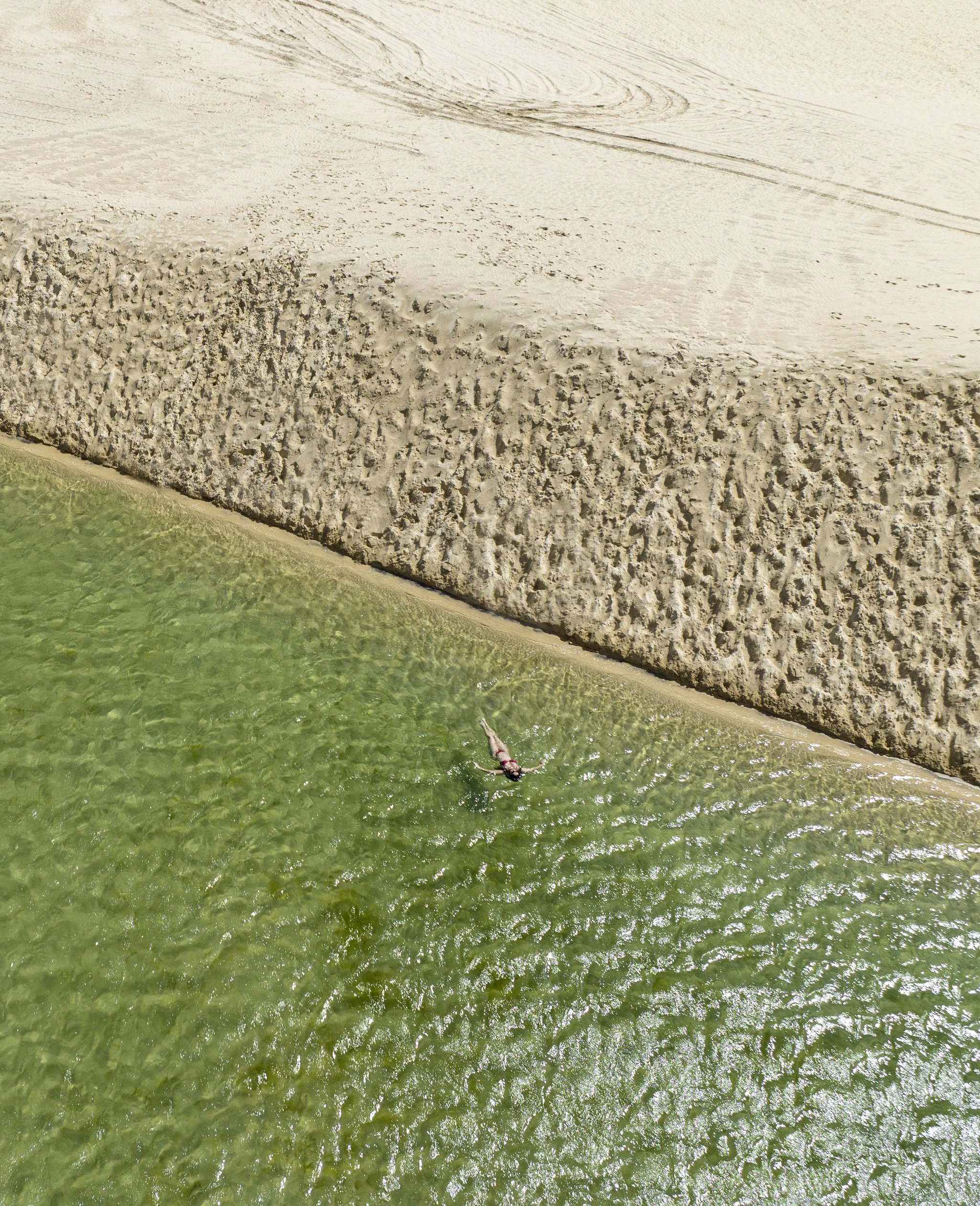Tiny against the dunes — one swimmer, infinite sand. Photo: Naeblys Aerial view of a lone swimmer floating in clear green water beside a vast sculpted sandbank on the Lençóis Maranhenses trek from Atins