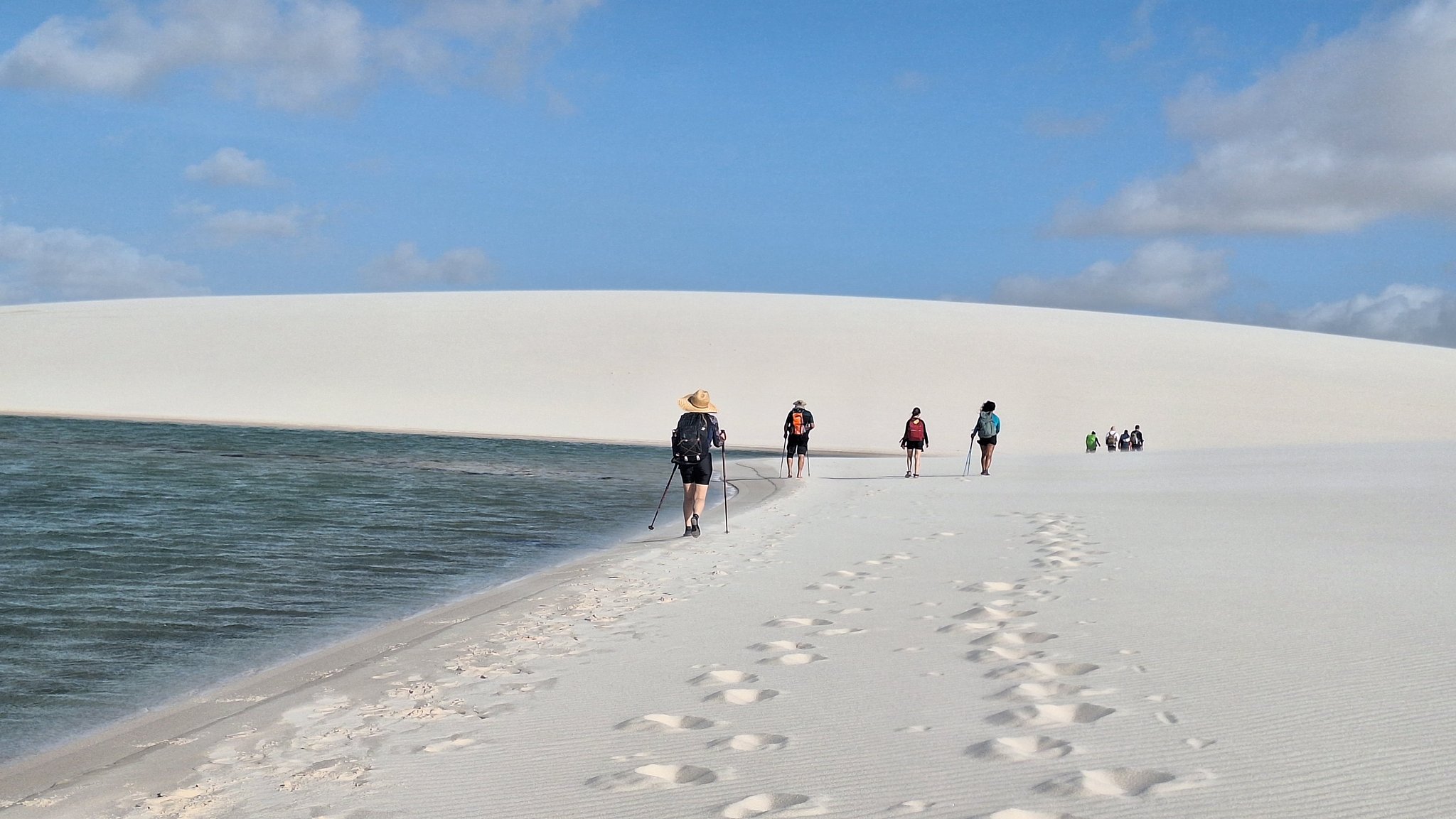 Sand ridge, dark lagoon, open sky — one foot in front of the other. A group of trekkers with backpacks and hiking poles walks along a narrow white sand ridge beside a dark-water lagoon during the Lençóis Maranhenses trek from Atins, with a sweeping dune crest meeting a blue sky above