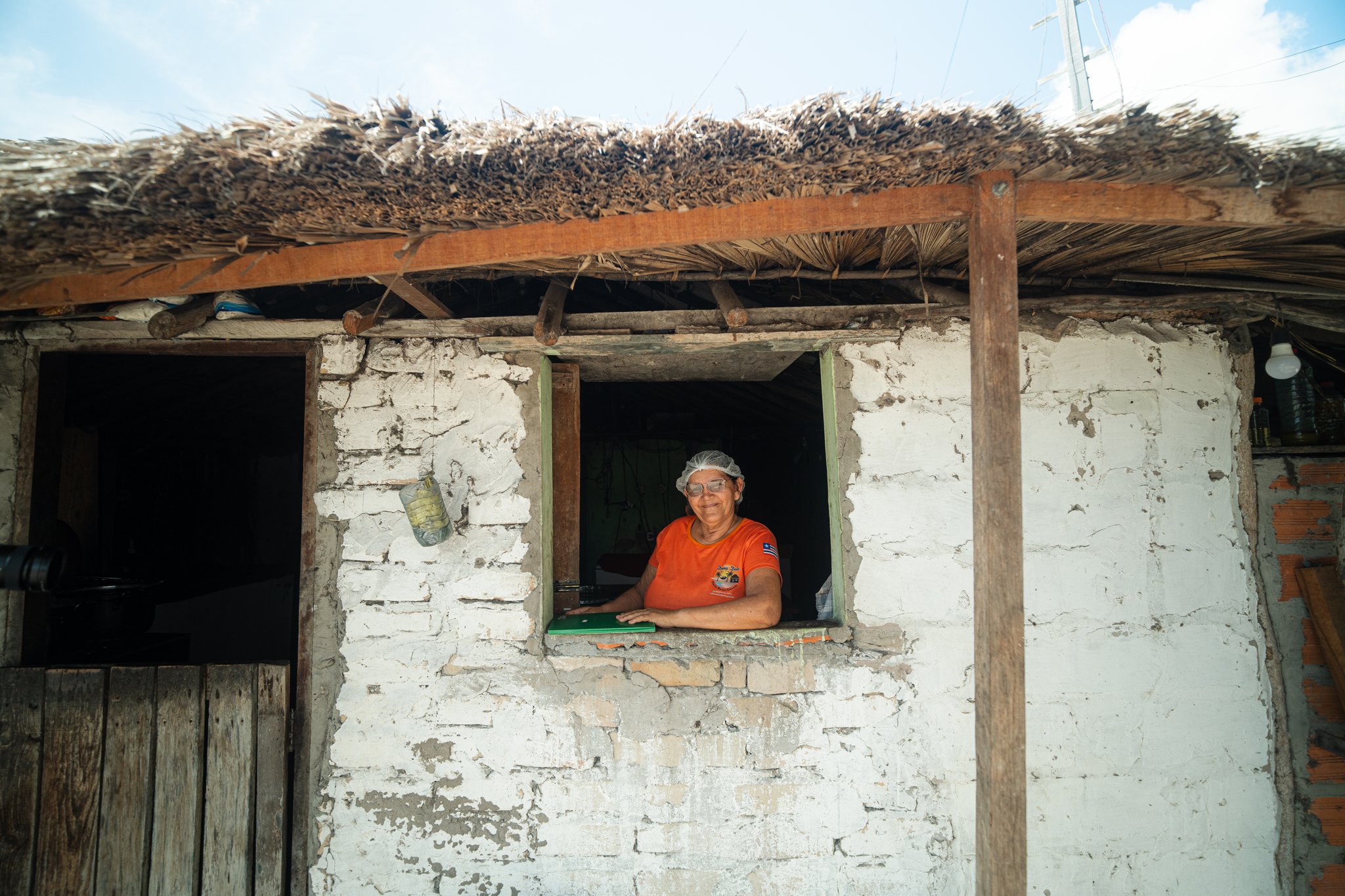 A warm face at the window — your host for the night in the dunes. A smiling woman wearing an orange shirt and hairnet leans out of the window of a rustic whitewashed brick house with a thatched palm roof, part of the Lençóis Maranhenses trek from Atins homestay experience
