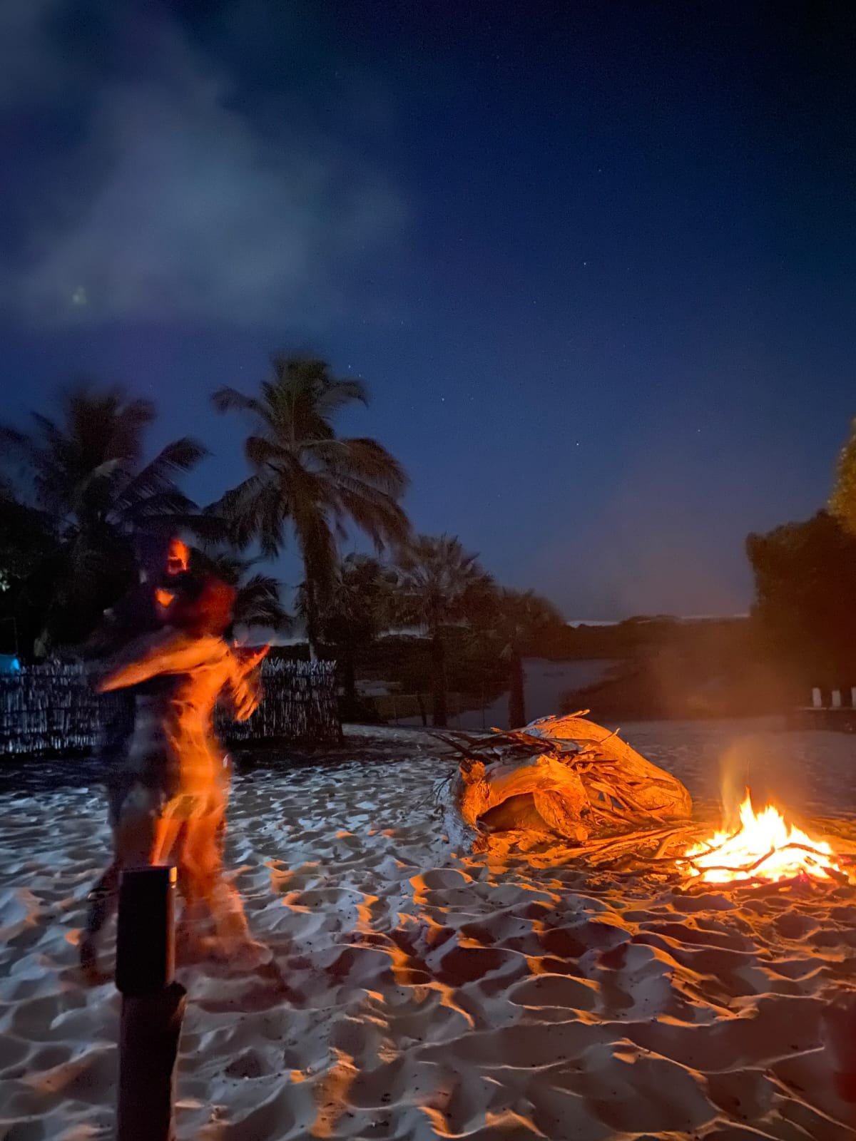 Firelight, soft sand, and a night that belongs entirely to you. A motion-blurred figure dances beside a crackling bonfire on a white-sand beach at night, palm trees silhouetted against a deep blue sky during a lencois maranhenses trek