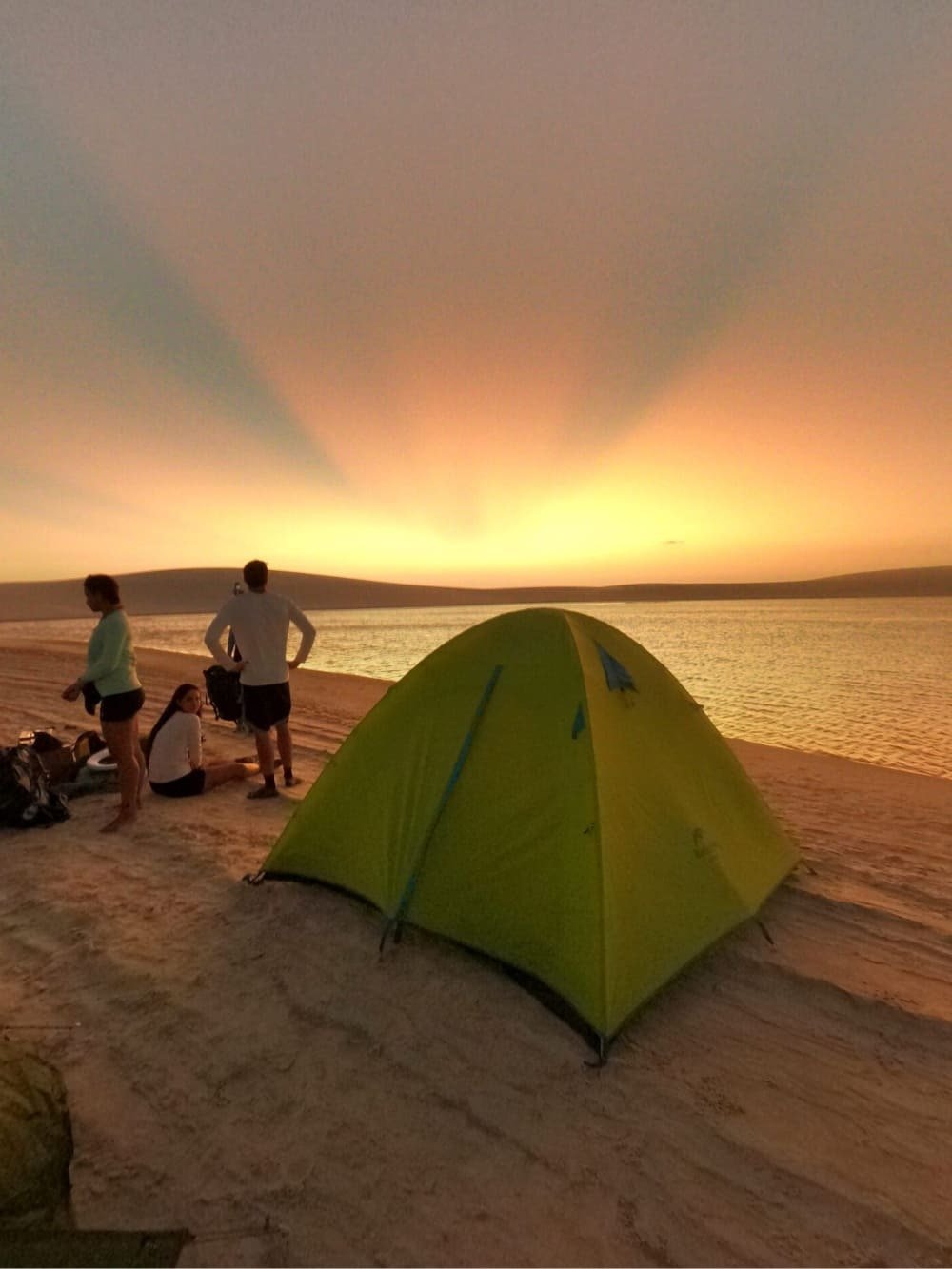 A green dome tent pitched on a sandy riverbank during a lencois maranhenses trek, with three travellers watching a vivid golden sunset over calm water and low dunes on the far shore