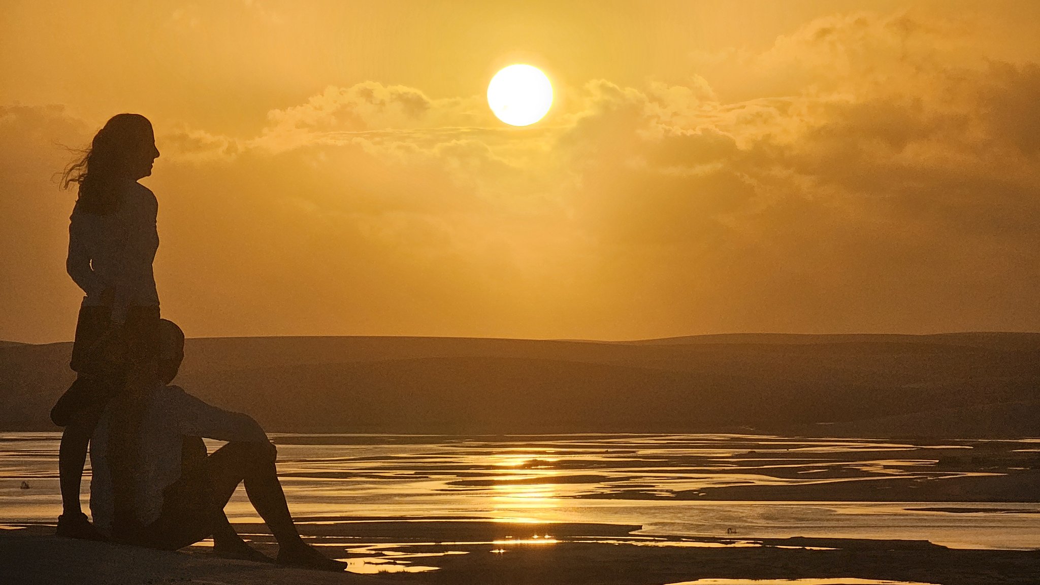 Still air, molten gold — the day ends at the edge of the dunes. Two silhouettes of travellers watching a golden sunset over the flat wetlands and dunes of a lençóis maranhenses trek, the sun burning bright through scattered clouds