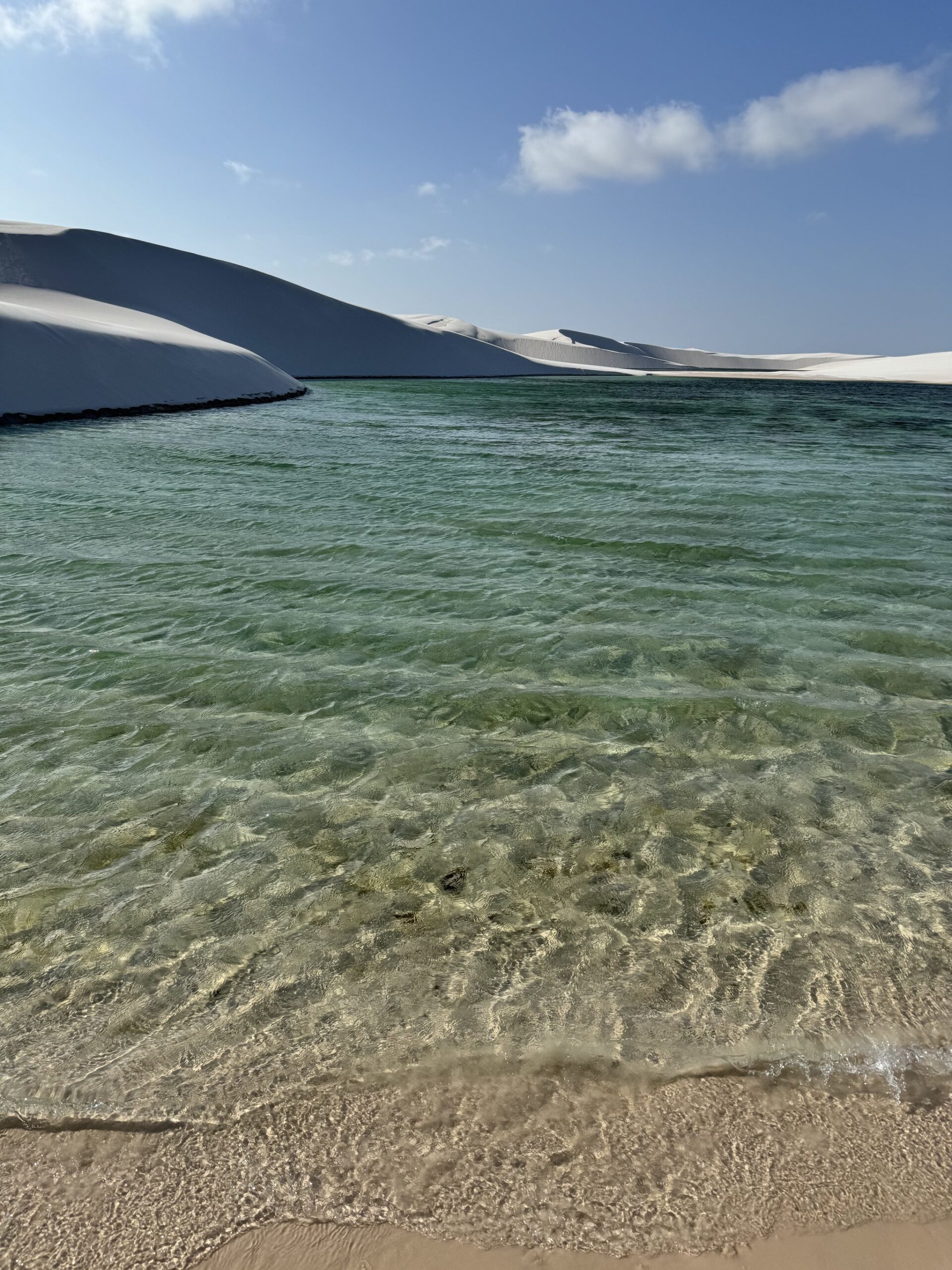 Turquoise water, white dunes — cooler than it looks, and cold enough to stop you mid-stride. Crystal-clear turquoise water of a freshwater lagoon meets white sand dunes on the Lençóis Maranhenses trek under a blue sky