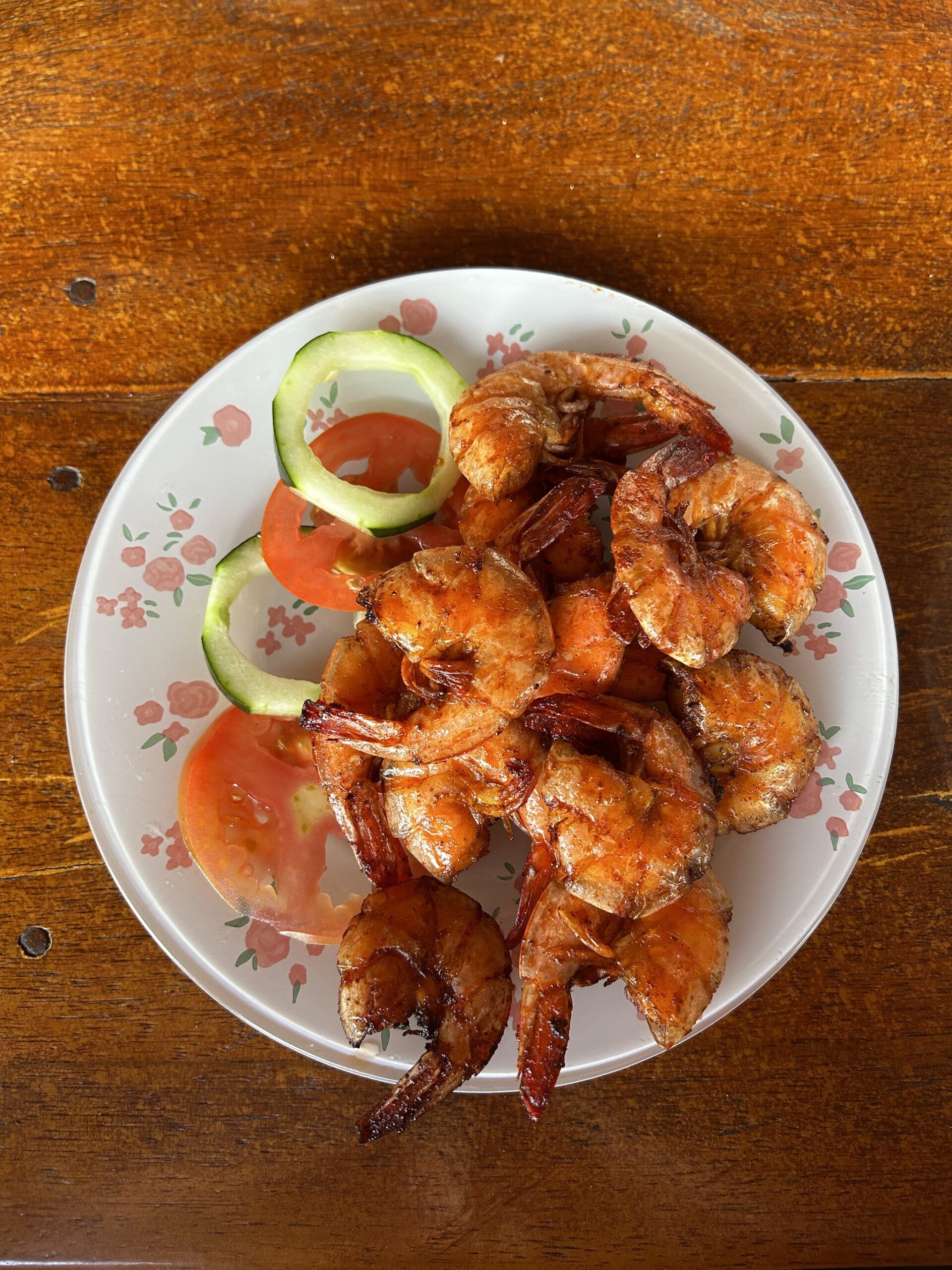 Fresh, charred, and simply perfect — seafood straight from the hands of your host family. A plate of golden grilled whole shrimp garnished with sliced tomato and cucumber rings, served on a floral-patterned dish on a wooden table during a lencois maranhenses trek homestay meal