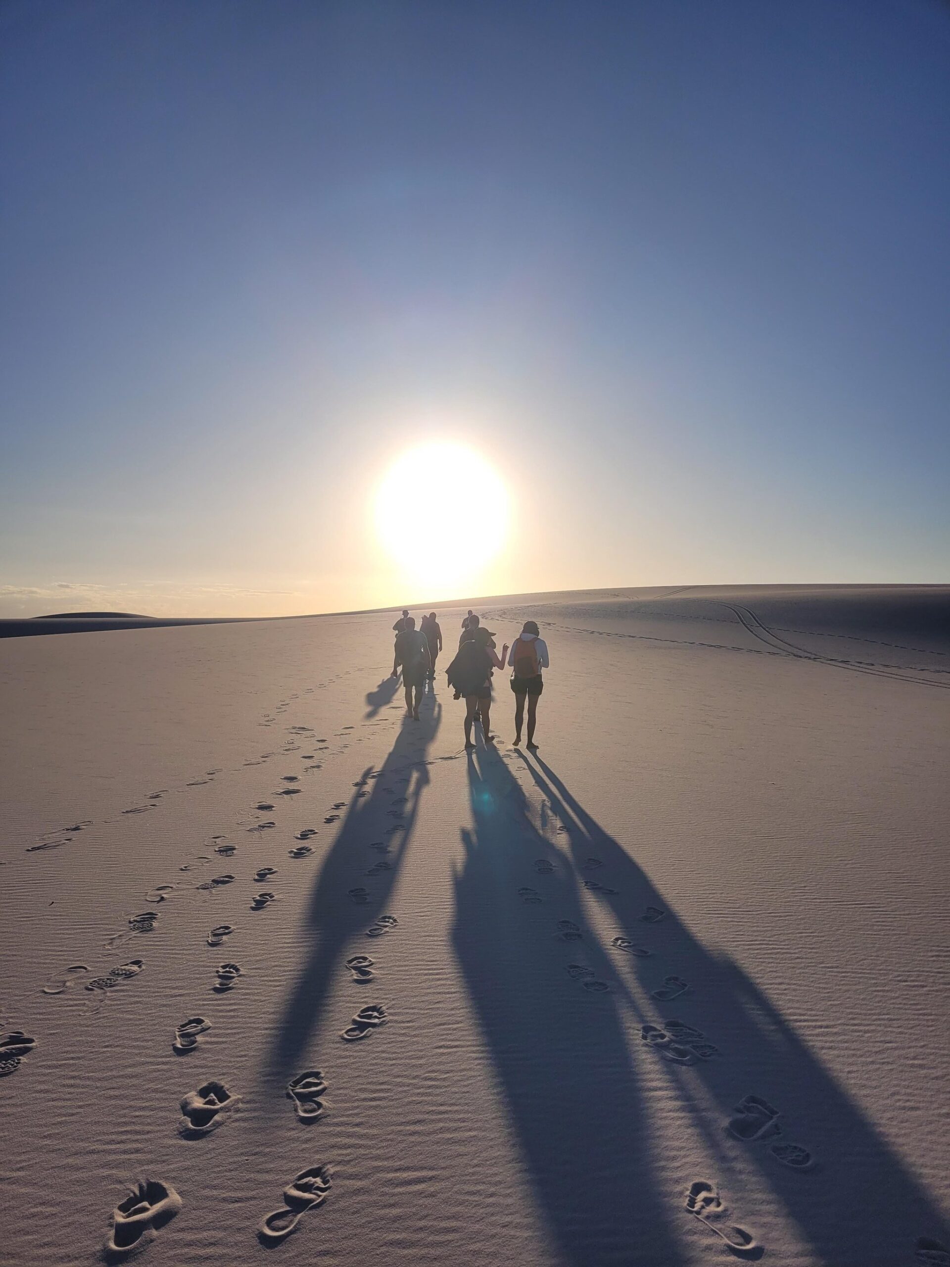 Six shadows, one blazing sun — the dunes swallow you whole. A group of trekkers walks toward a blazing sun on the lencois maranhenses trek, their long shadows and barefoot footprints stretching across pale white sand dunes under a clear blue sky