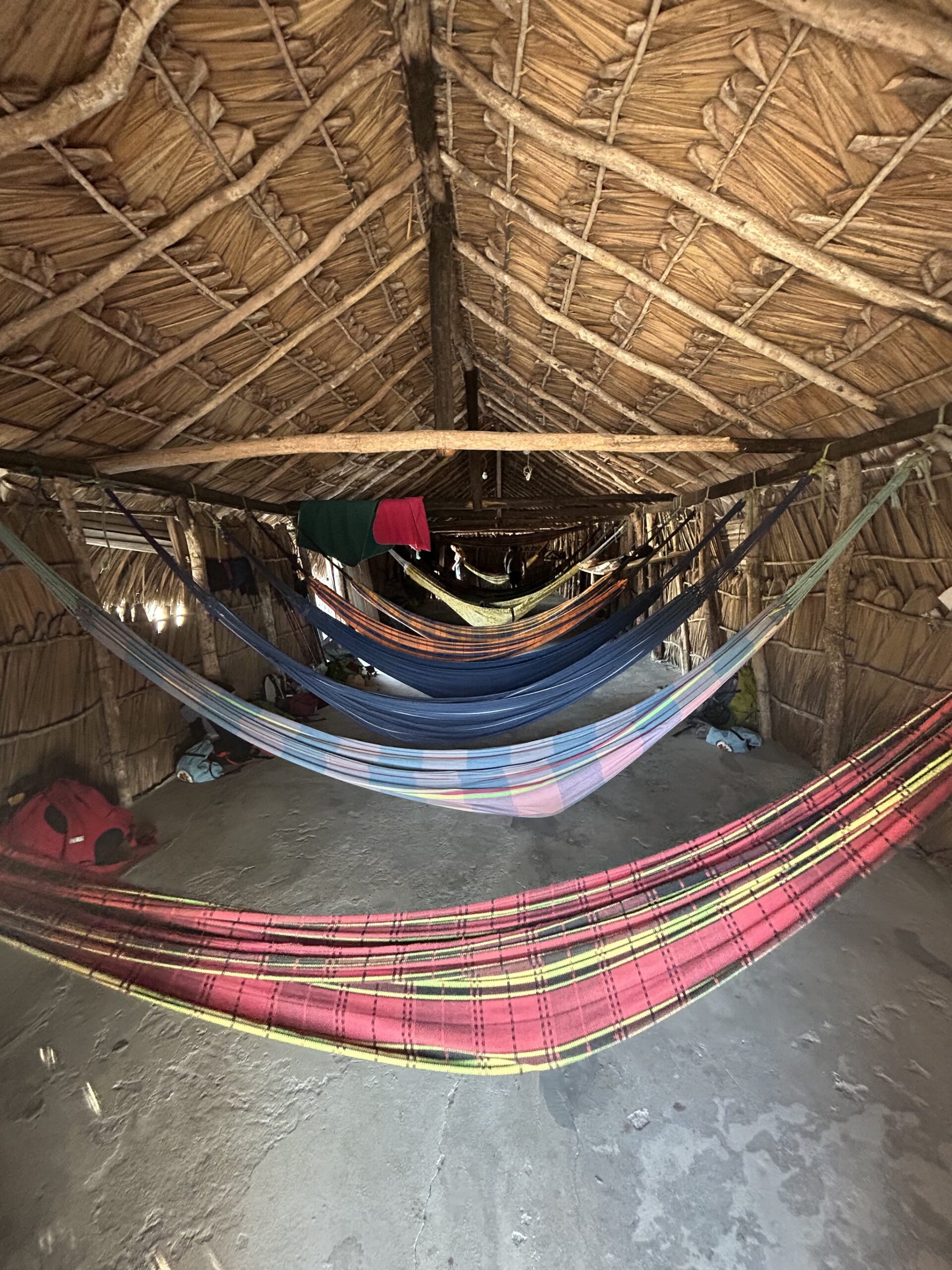 Rows of colourful hammocks suspended inside a rustic thatched-roof shelter on the Lençóis Maranhenses trek, with travellers' bags resting on the earthen floor below