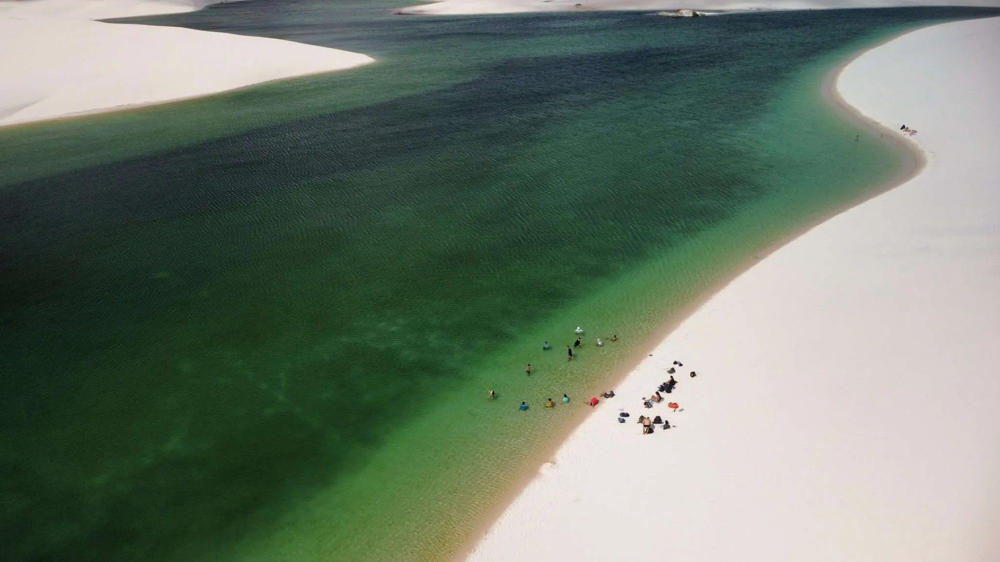 Aerial view of swimmers wading along the curved white sand shore of a large emerald and deep-blue freshwater lagoon during a lencois maranhenses trek