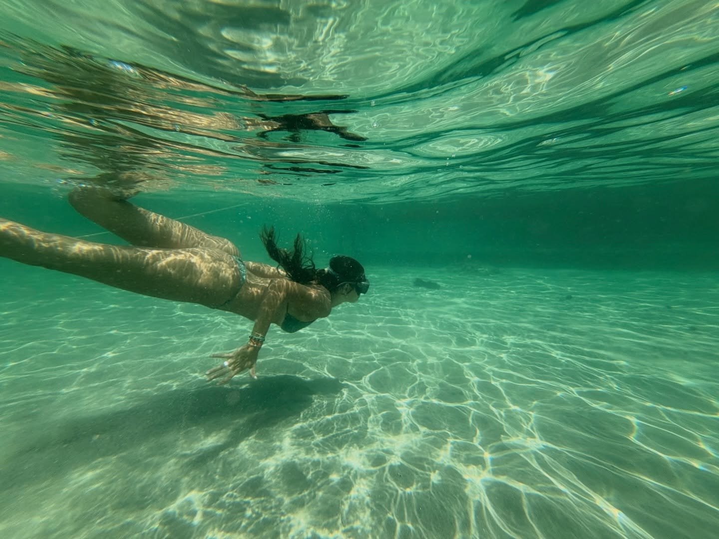 Weightless in water so clear you can see every ripple on the sand below. A woman free-diving through crystal-clear turquoise water above a sandy floor during a lencois maranhenses trek lagoon stop