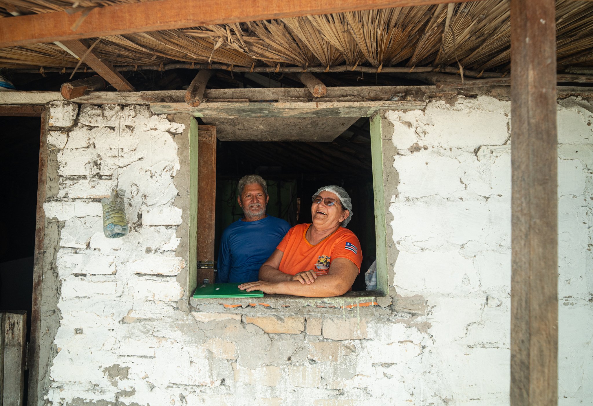 Laughter through a whitewashed window — the real heart of the oasis. Two local residents — a man in a blue shirt and a woman in an orange shirt wearing a hair net — lean out of a whitewashed stone window with a thatched roof overhead, laughing warmly on a lençois maranhenses trek homestay stop