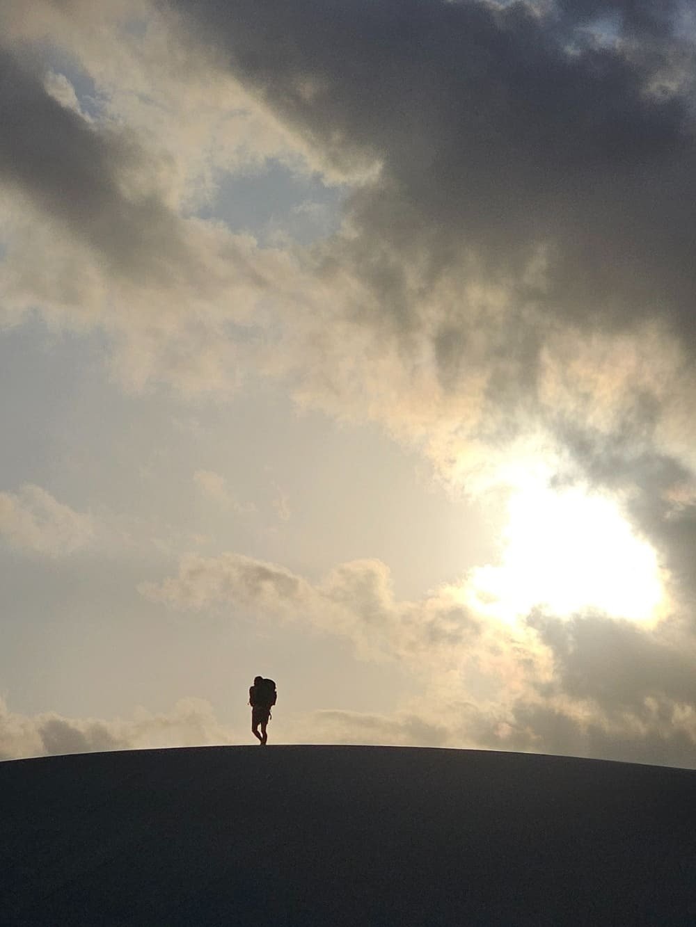 Small figure, boundless sky — the dunes put everything in perspective. A lone backpacker silhouetted on the crest of a vast sand dune during the Lençóis Maranhenses trek, beneath a dramatic sky with the sun breaking through clouds