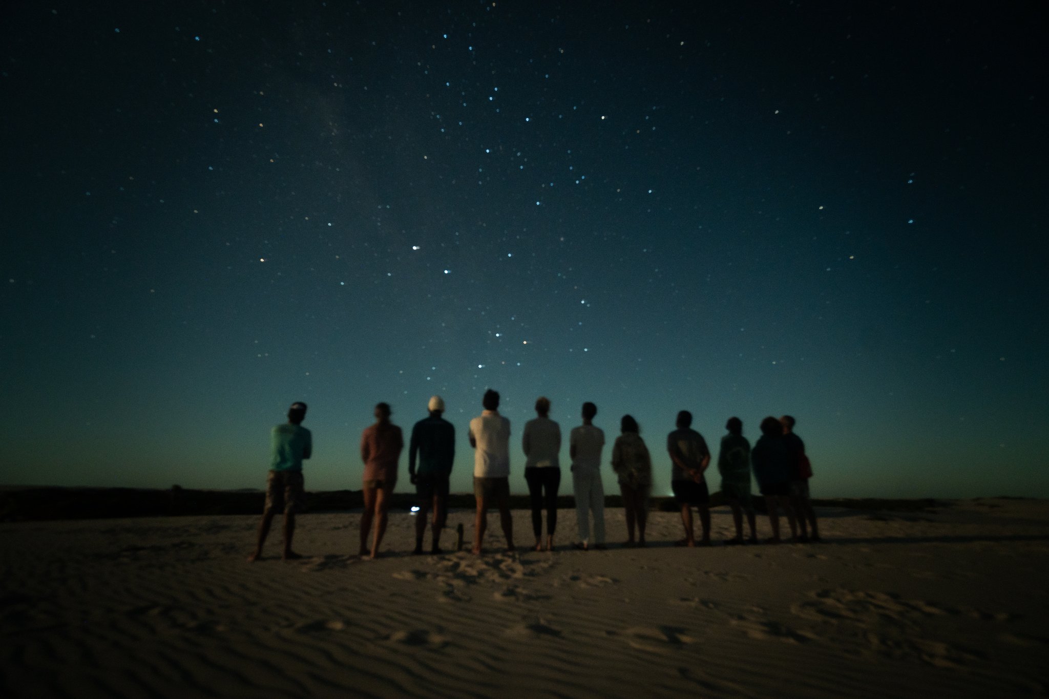 Ten travellers, a boundless sky, and more stars than you can count. A group of ten travellers stands in silhouette on open sand dunes beneath a vast star-filled sky during a lencois maranhenses trek night stop