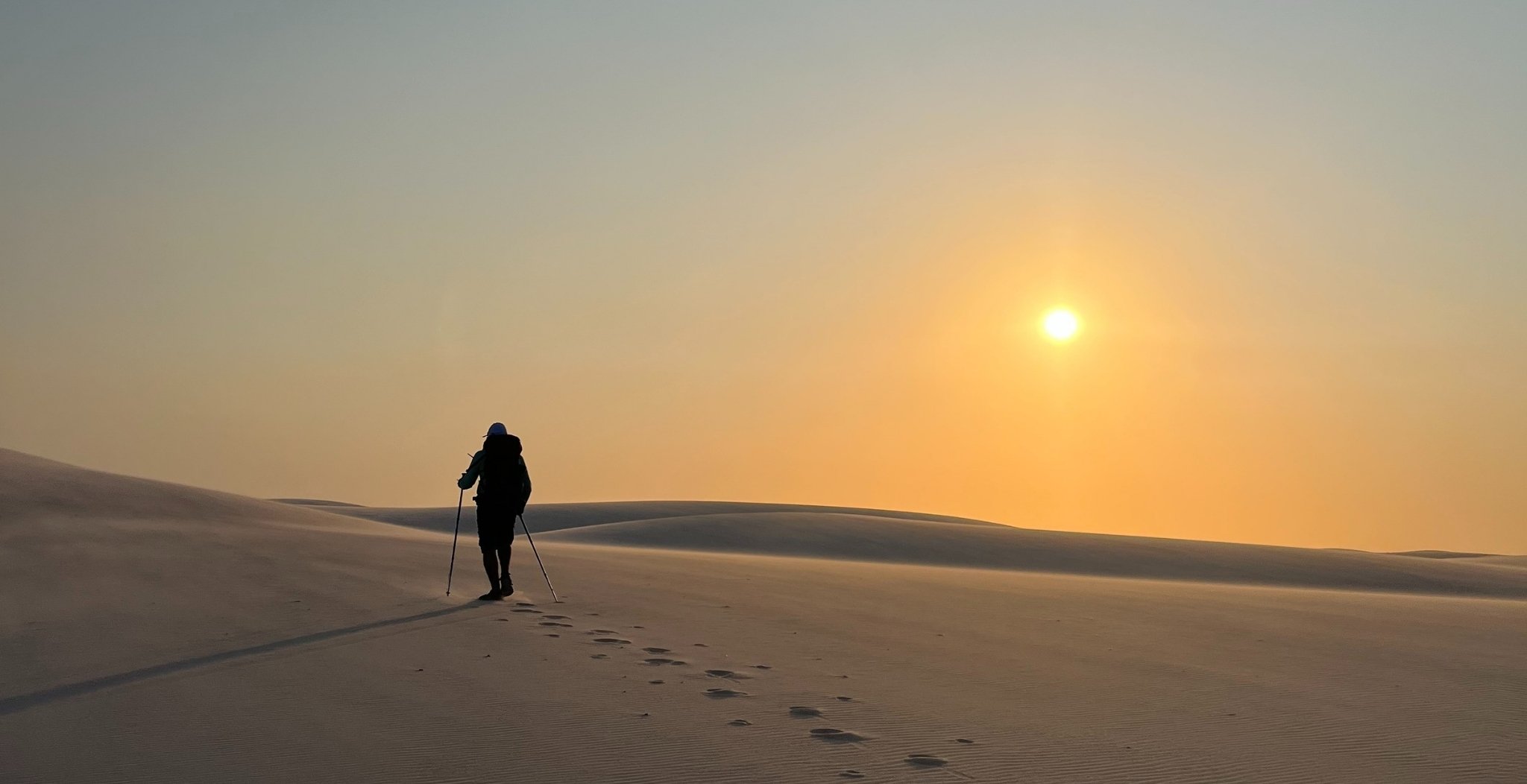 A lone trekker with hiking poles walks up a white sand dune toward the rising sun on a lencois maranhenses trek, footprints trailing behind across the smooth, wind-rippled sand