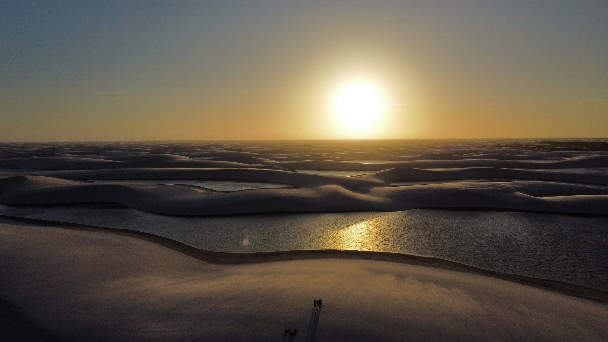 Two silhouettes, an infinite dunefield, and a sun that turns every lagoon to gold. Aerial view of the lencois maranhenses trek at golden-hour sunset, with vast white sand dunes casting long shadows over scattered freshwater lagoons reflecting the sun, and two tiny silhouetted figures standing on the dune foreground