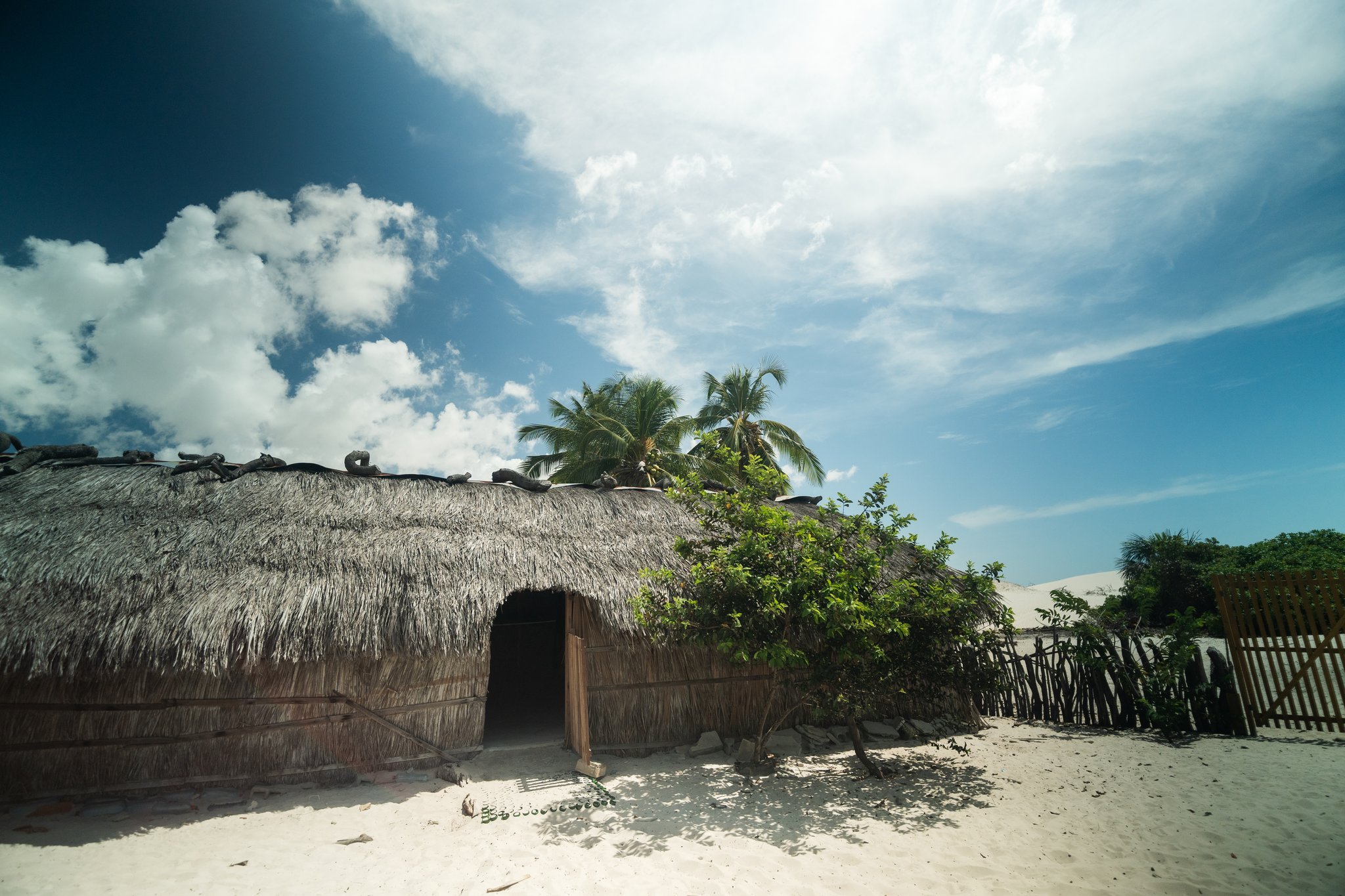A traditional thatched-roof dwelling on white sand with palm trees behind it, part of a lencois maranhenses trek oasis community homestay