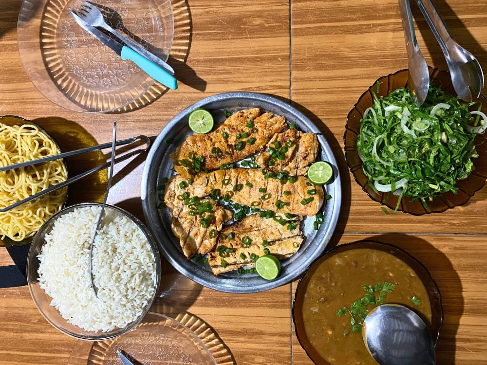 Generous, colourful, and cooked with pride — a family table inside the park. Overhead view of a shared meal table on the lencois maranhenses trek, featuring grilled seasoned fish fillets topped with spring onion and lime, white rice, noodles, a green herb salad, and a thick brown bean stew