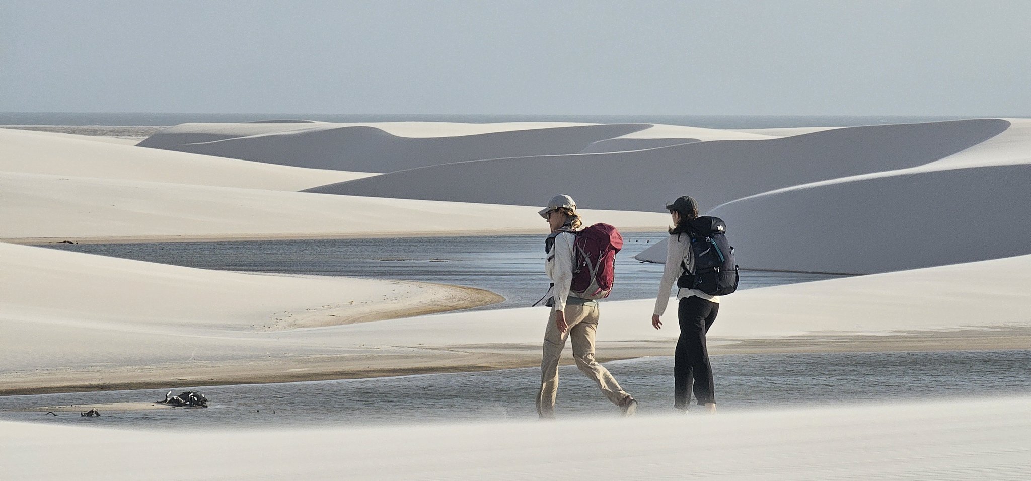 Two packs, two footprints, and a landscape that feels like the edge of the world. Two backpackers walk side by side across the white sand dunes of the lencois maranhenses trek, with a shallow freshwater lagoon and sweeping dune ridges stretching to the horizon behind them