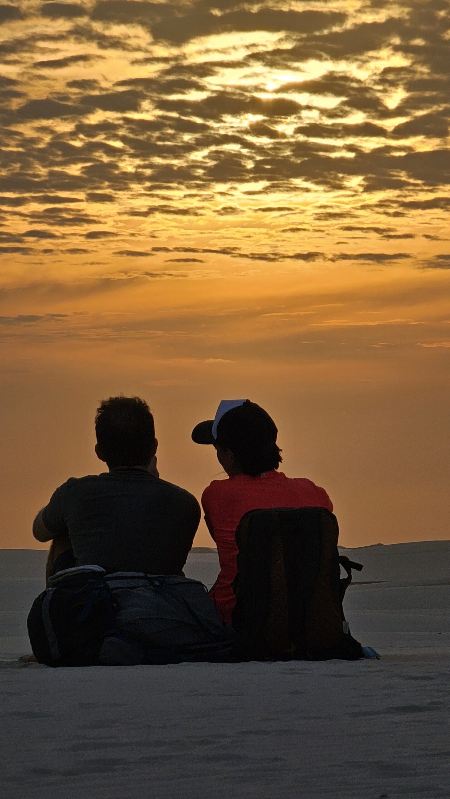 Side by side, nowhere else to be. Two travelers sit silhouetted on white sand dunes during a Lençóis Maranhenses trekking sunset, facing a vivid golden sky filled with scattered clouds