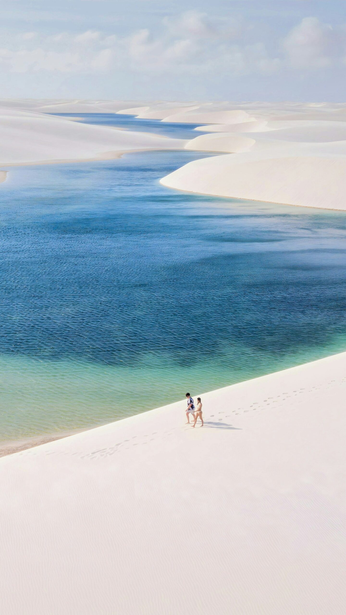 Two travelers walking barefoot along a sweeping white sand dune beside a vast turquoise lagoon during a Lençóis Maranhenses trekking expedition, with more dunes and blue water stretching to the horizon