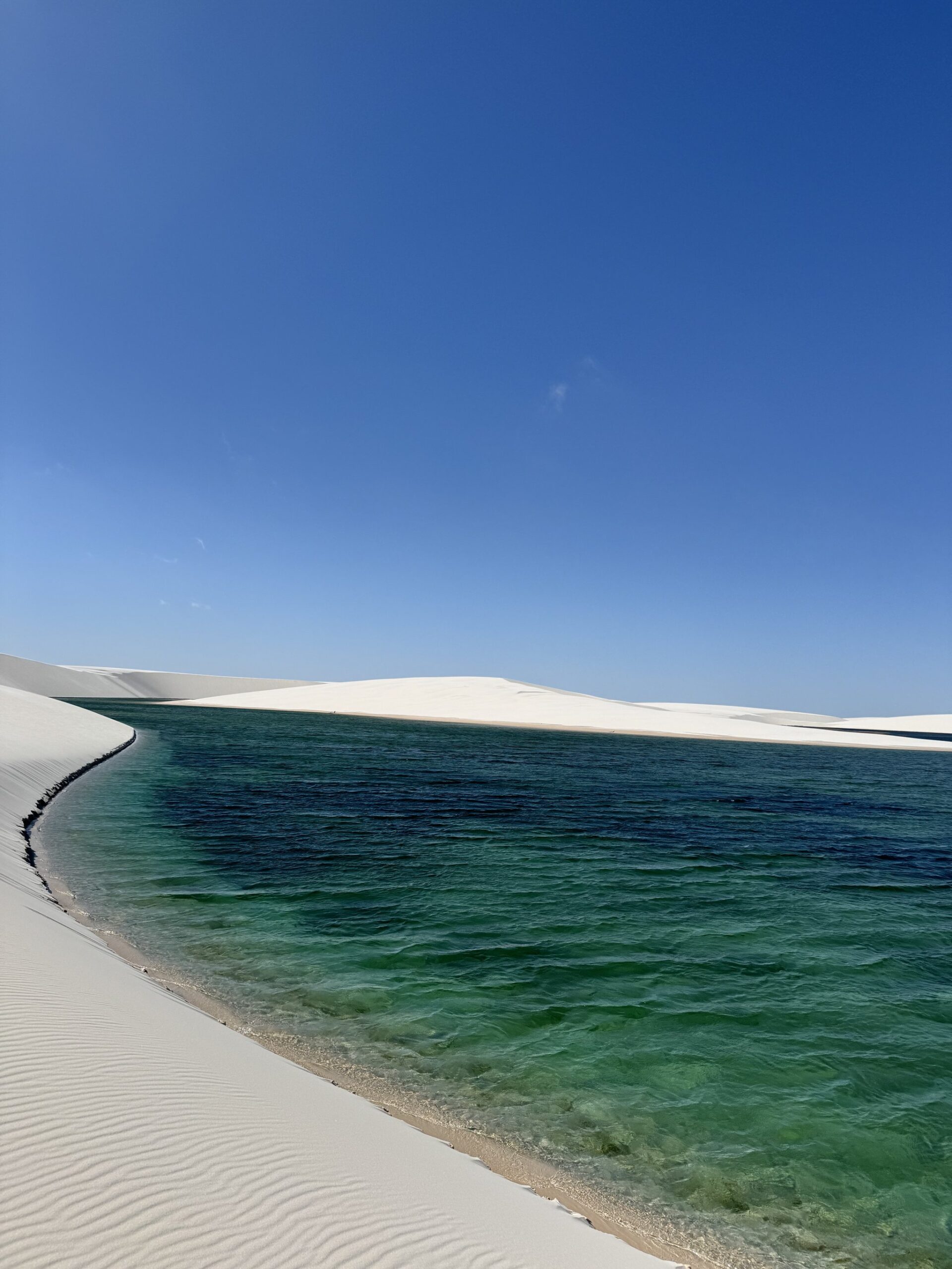 Pure emerald, pure white — a lagoon so still it looks painted. Crystal-clear emerald lagoon nestled against sweeping white sand dunes under a deep blue sky in Lençóis Maranhenses trekking country