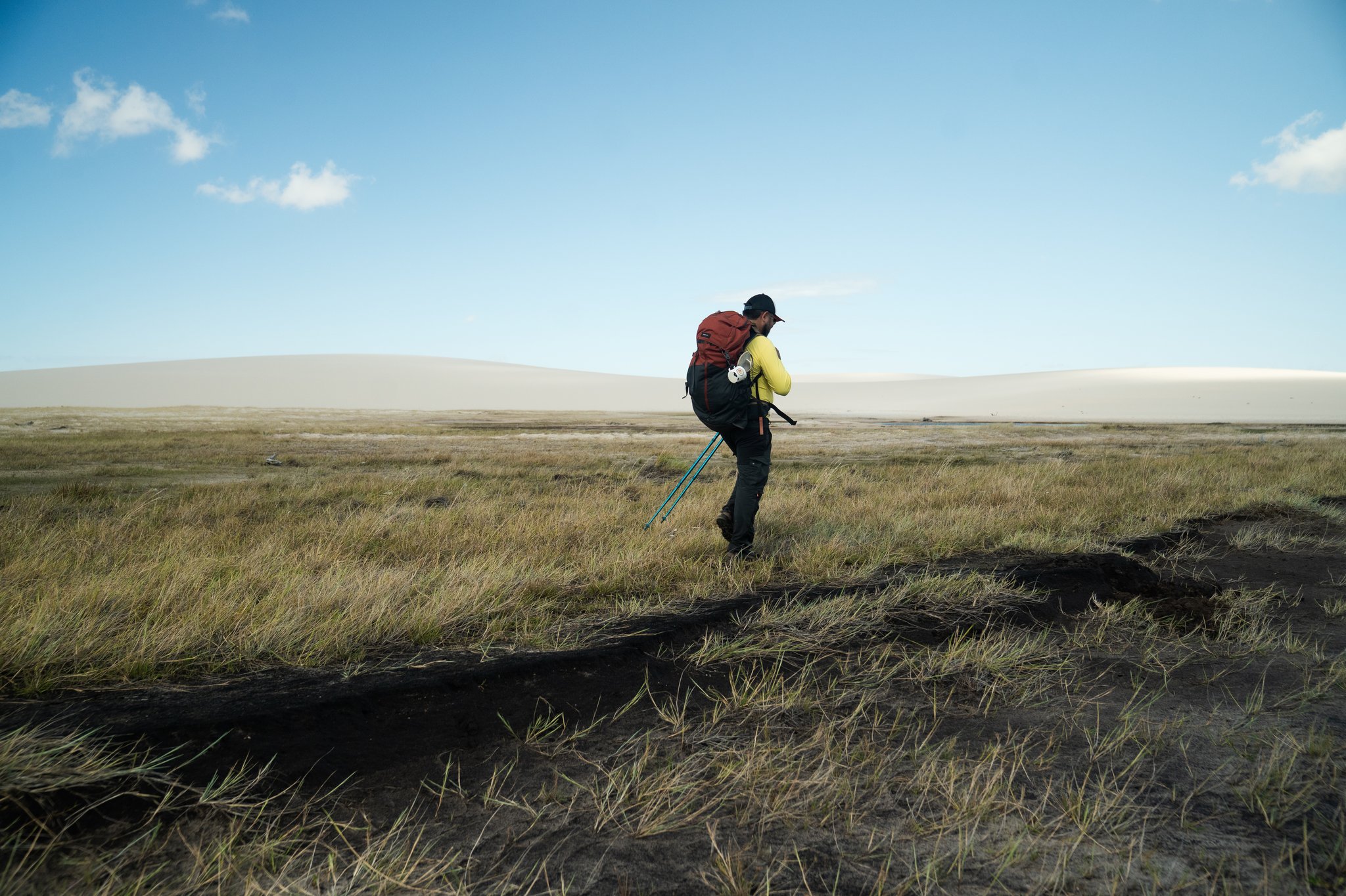 A trekker with a red backpack and trekking poles crosses a windswept grassy plain during a Lençóis Maranhenses trekking expedition, with pale sand dunes rising on the horizon under a wide blue sky