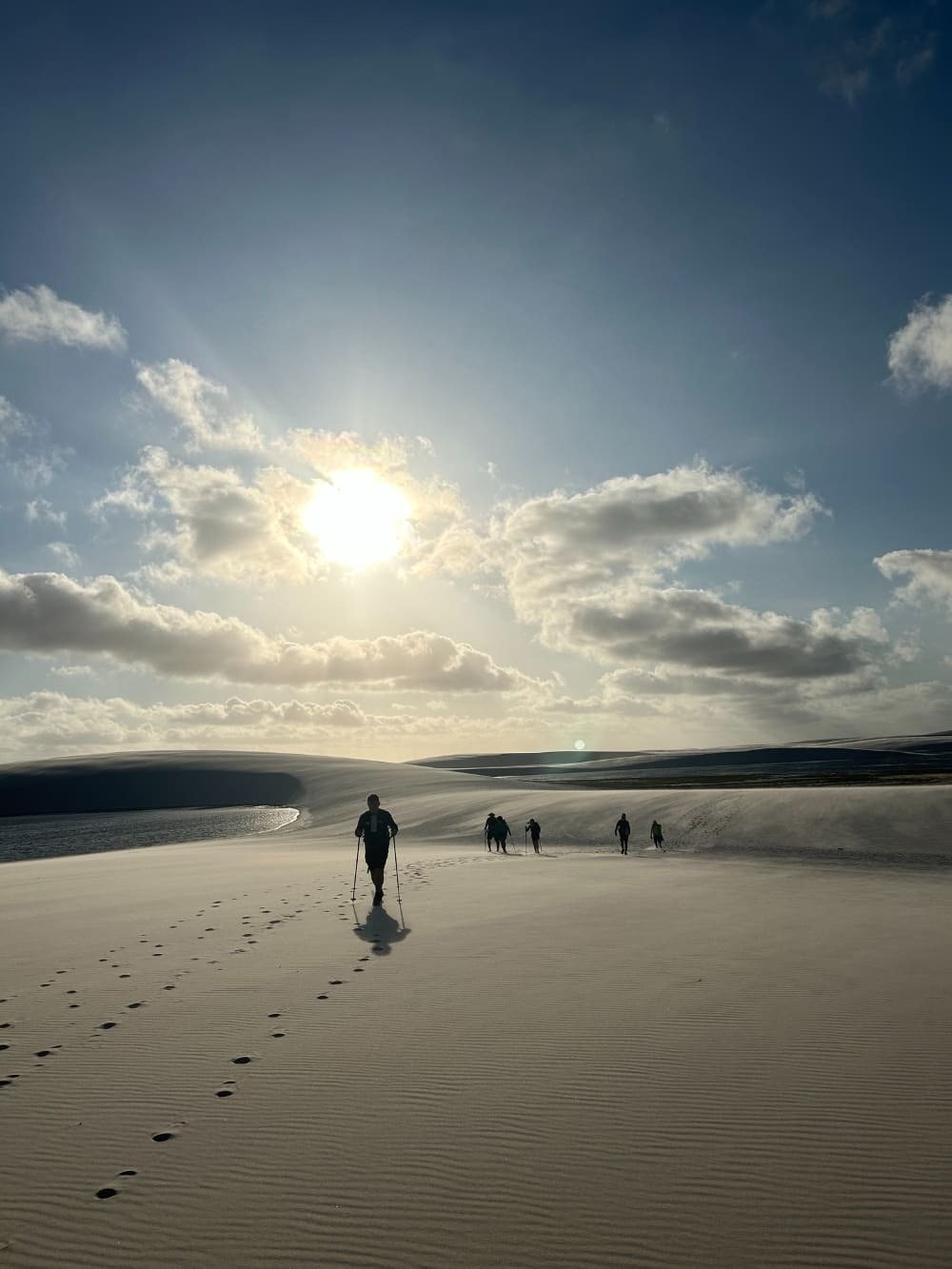 A group of trekkers walks across vast white sand dunes under a bright sun during a Lençóis Maranhenses trekking expedition, with a dark lagoon glinting to the left and rolling dunes stretching to the horizon