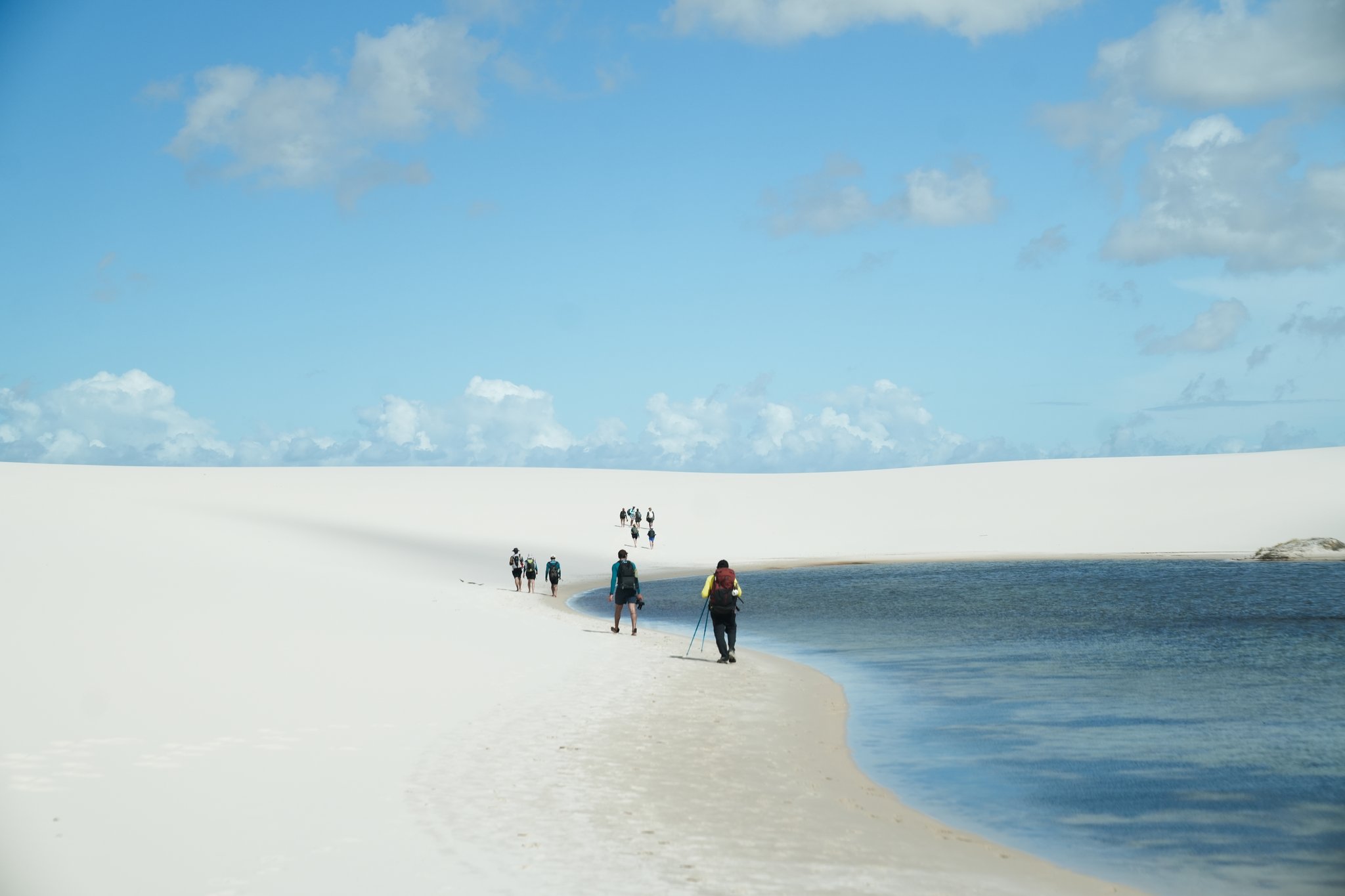 Trekkers stretched thin across the dune ridge, lagoon inches to the right. A line of backpackers walking along a narrow white sandbar beside a clear blue lagoon during a Lençóis Maranhenses trekking crossing, under a bright sky with scattered clouds