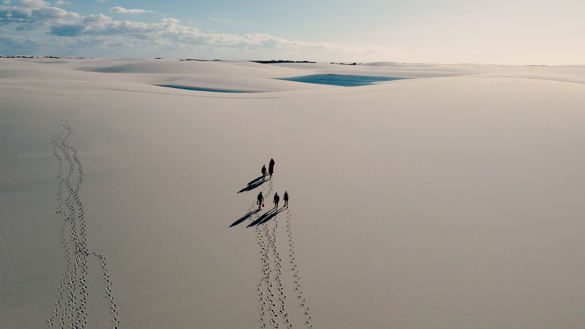 Five tiny figures, endless white — the dunes swallow everything but footprints. Aerial view of five trekkers casting long shadows across vast white sand dunes during a Lençóis Maranhenses trekking experience, with blue rain-filled lagoons visible in the distance