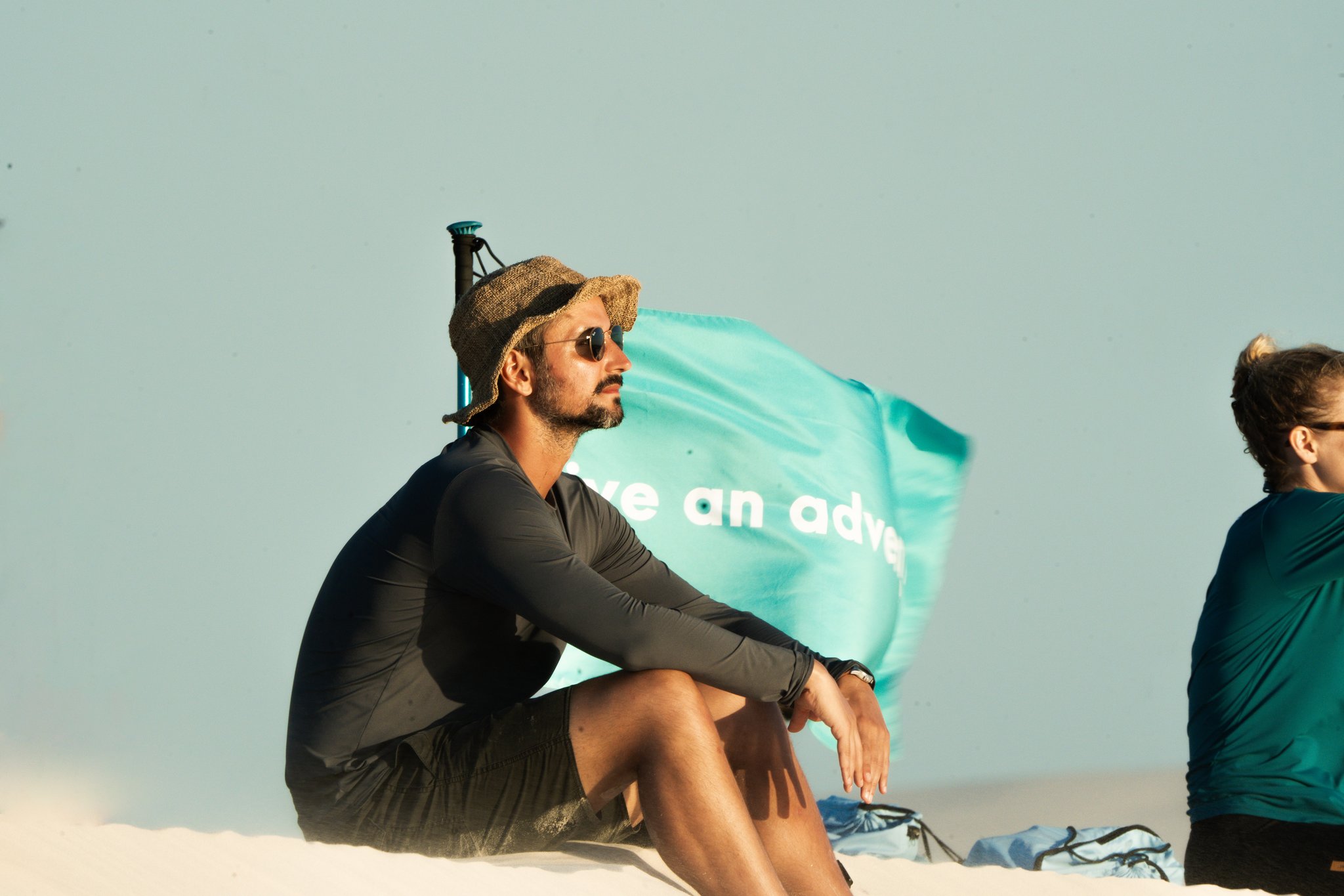 A quiet moment between miles — hat, shades, and endless white sand. A man in a straw hat and sunglasses sits on white sand dunes during a Lençóis Maranhenses trekking adventure, a turquoise flag billowing behind him in the wind