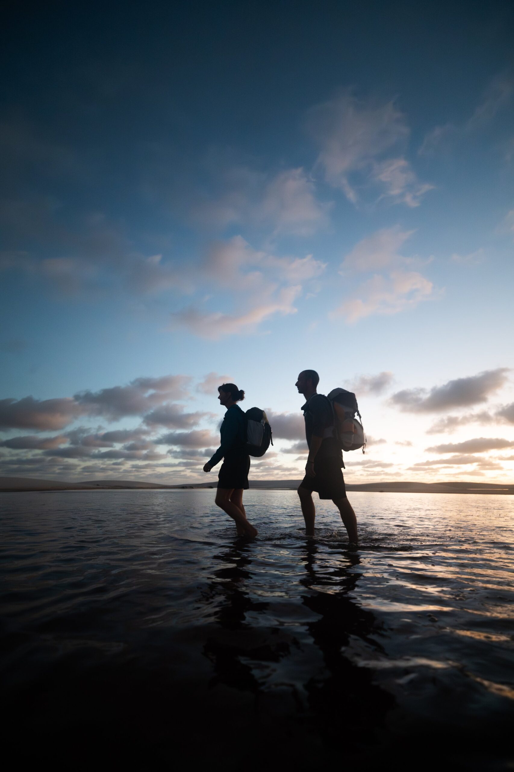 Barefoot, backpacks on, water underfoot — the trek ends as it lived. Two trekkers with backpacks silhouetted against a twilight sky as they wade barefoot through shallow water during a Lençóis Maranhenses trekking crossing