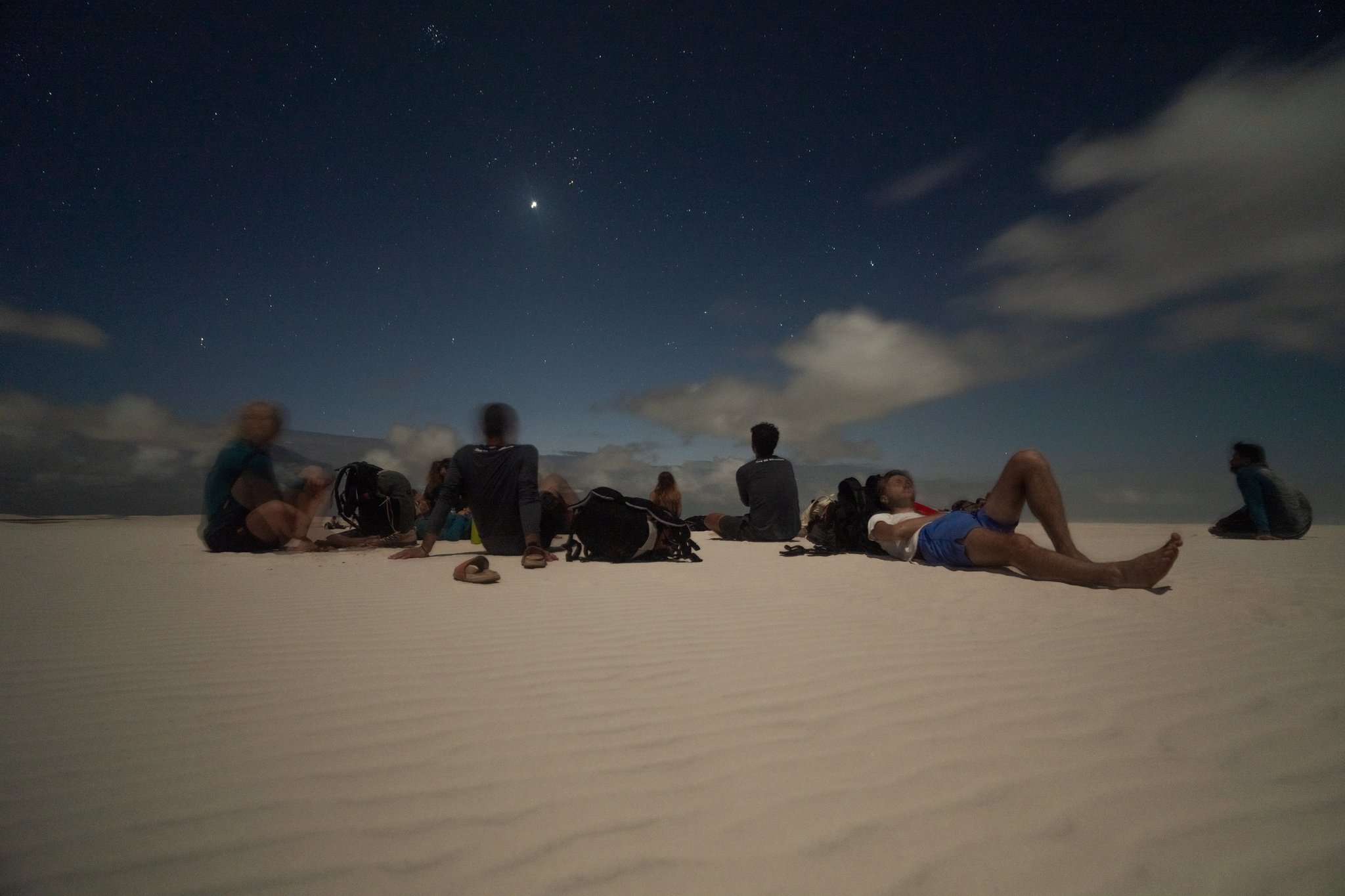Stars overhead, cool sand below — a night on the dunes that stays with you. A group of travelers lying and sitting on white sand dunes at night during a Lençóis Maranhenses trekking experience, gazing up at a star-filled sky with scattered clouds
