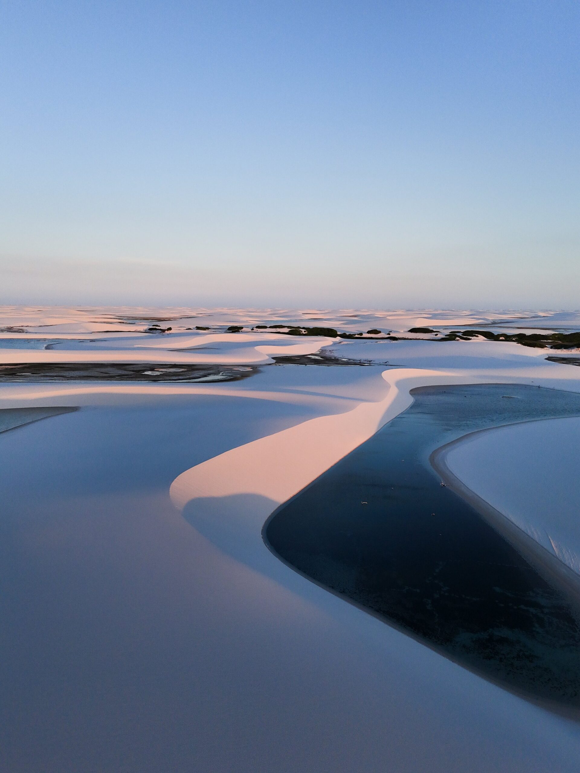 Pink light spills across the dunes, turning every lagoon into a mirror. Aerial view of white sand dunes and dark rain-filled lagoons glowing pink and blue at sunset during a Lençóis Maranhenses trekking crossing