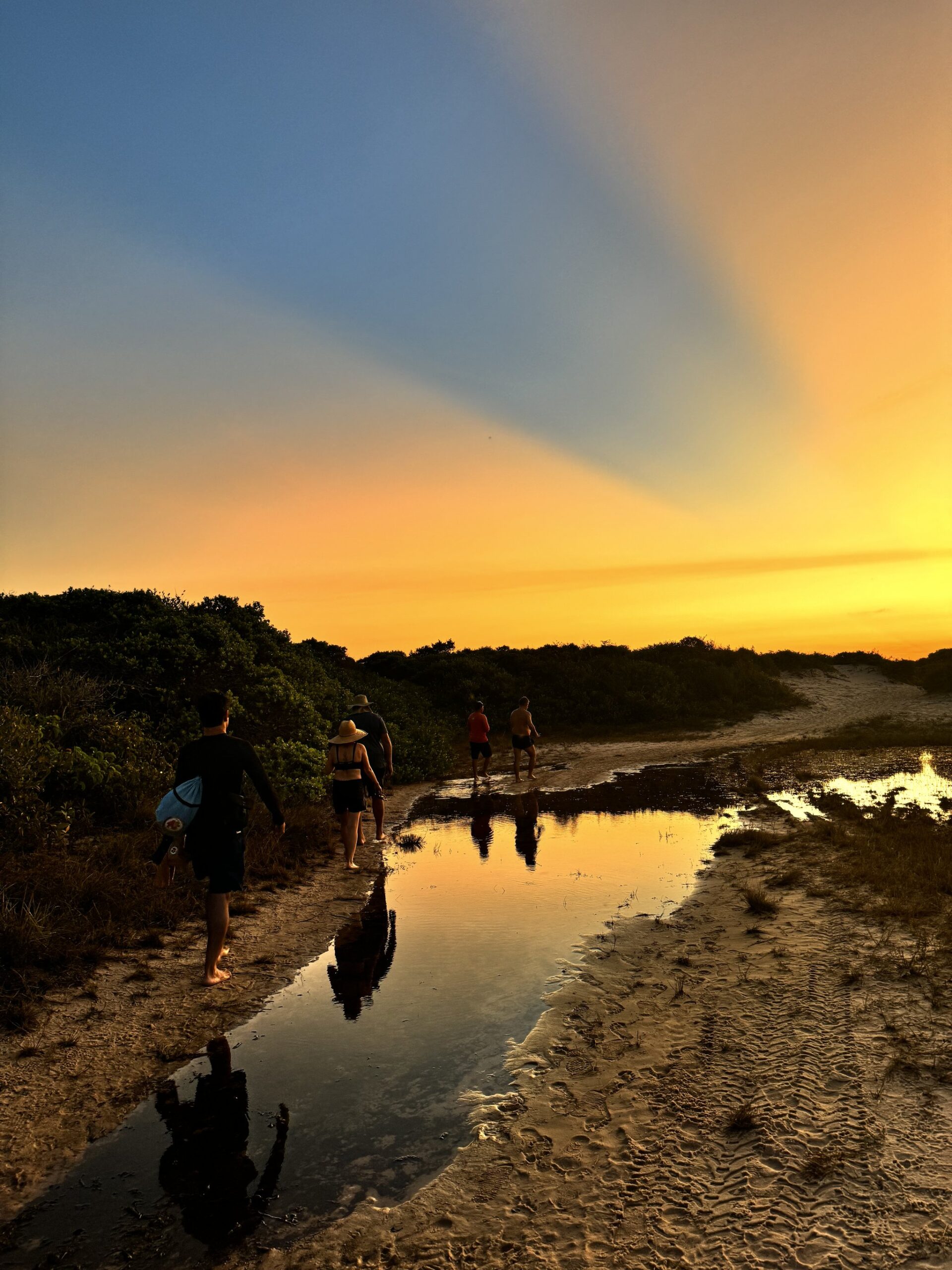Golden hour on the trail — the sky and the puddles agree. A group of trekkers walks barefoot along a sandy coastal path at golden hour, their silhouettes reflected in shallow standing water during a Lençóis Maranhenses trekking expedition, with low coastal scrub and a blazing orange sky ahead