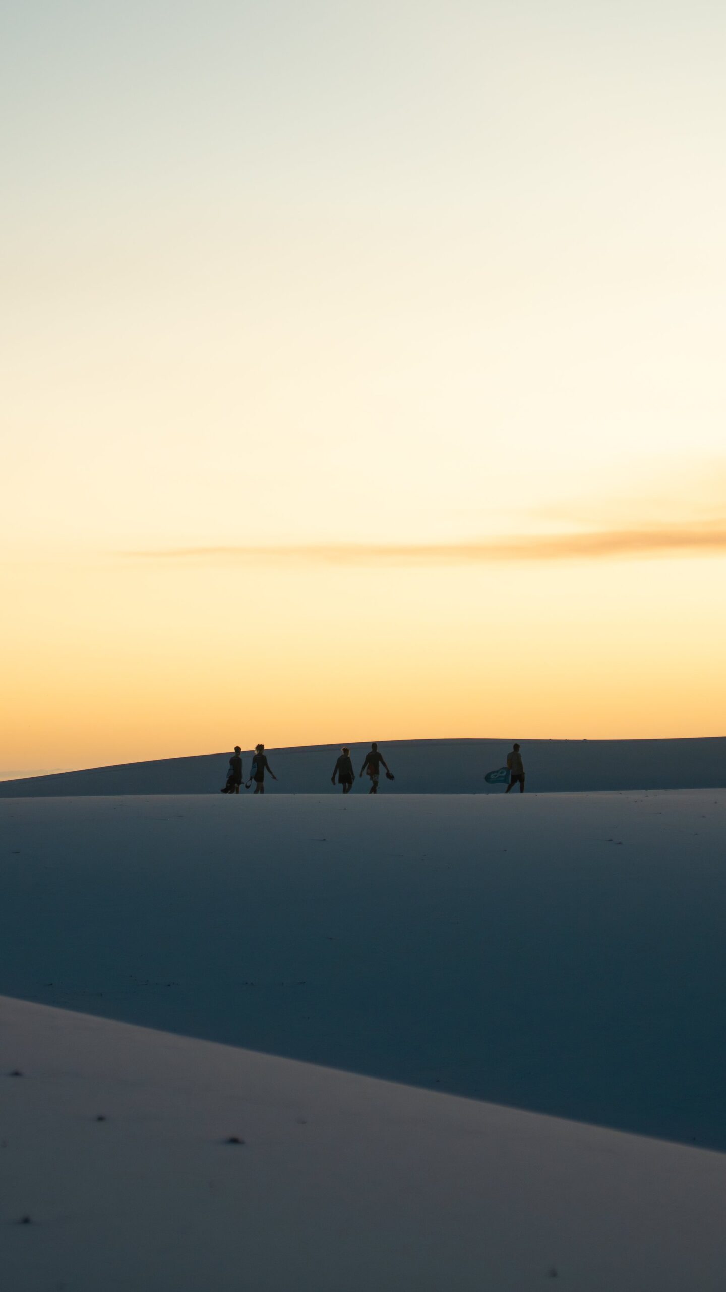Carrying boards, chasing the last light — the dunes belong to them now. Five silhouettes carrying sandboards walk along the crest of a white sand dune during a Lençóis Maranhenses trekking experience, set against a warm golden sunset sky