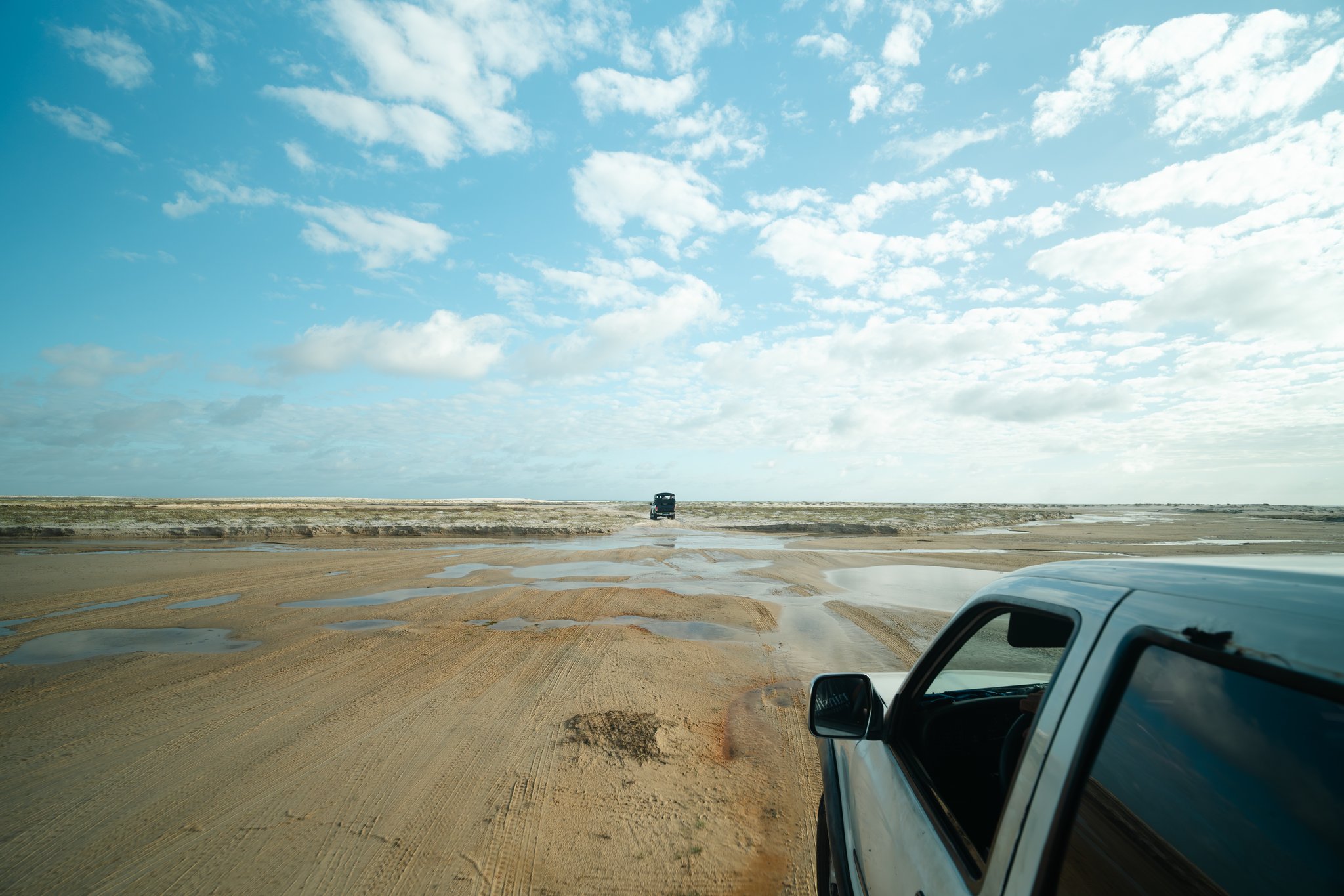 Puddles mirror the sky — the only road is the one you make. Two 4x4 vehicles crossing a vast wet sandy flat under a wide blue sky on a Lençóis Maranhenses trip, with shallow puddles reflecting light across the trackless terrain