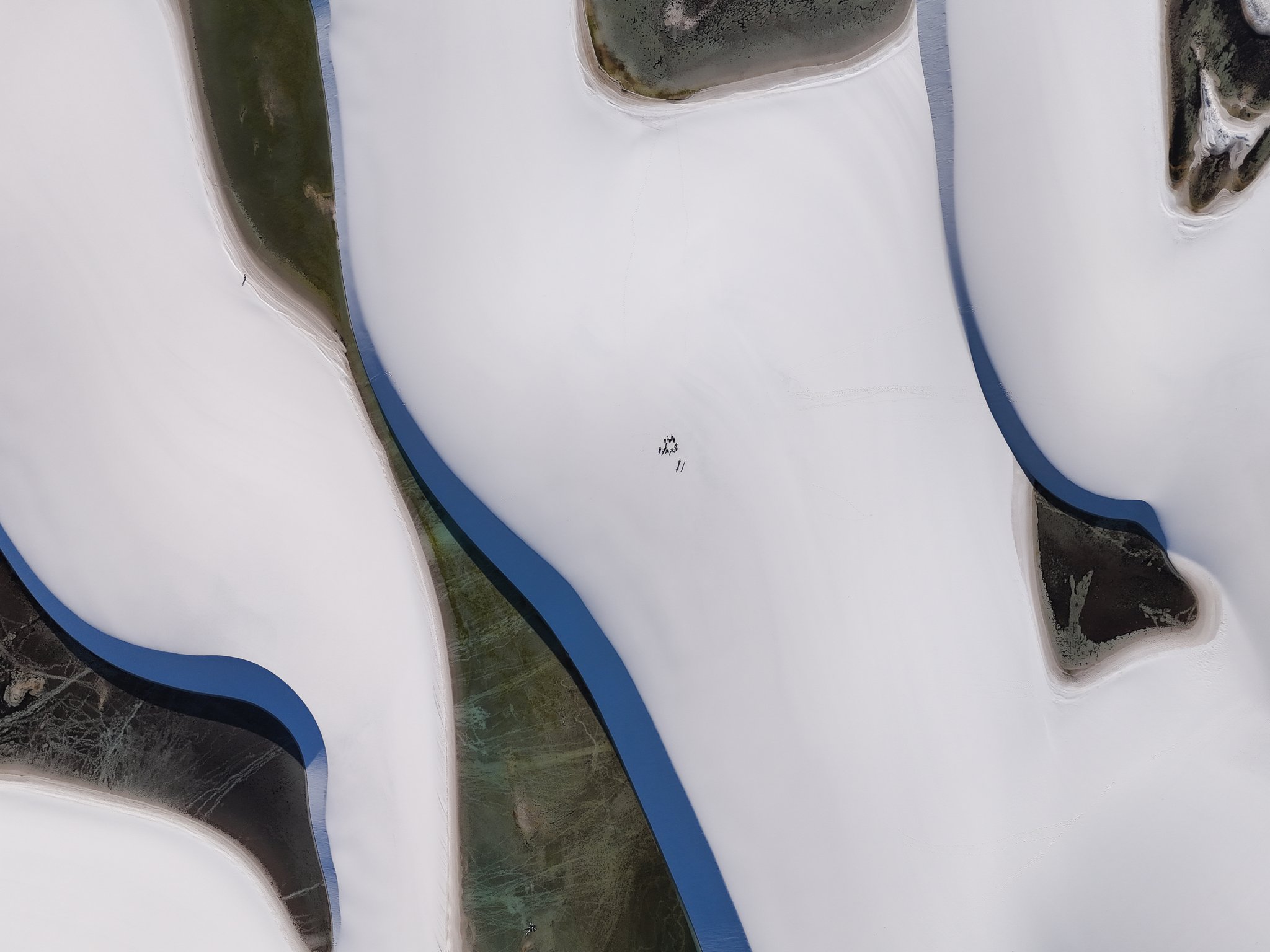 Aerial top-down view of vast white sand dunes on a Lençóis Maranhenses trip, with dark tannin-stained water channels weaving between the dune ridges and tiny figures visible at the center of the frame