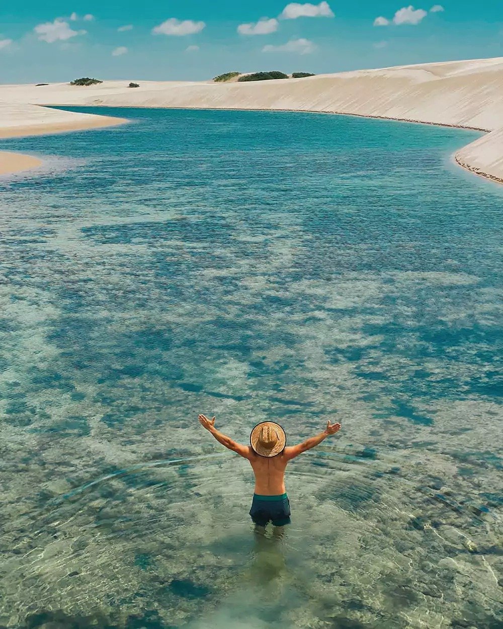 Arms wide open, water like glass — this is what the dunes reward. A traveler standing waist-deep in a crystal-clear turquoise lagoon on a Lençóis Maranhenses trip, arms raised wide, with sweeping white sand dunes stretching into the distance under a bright blue sky