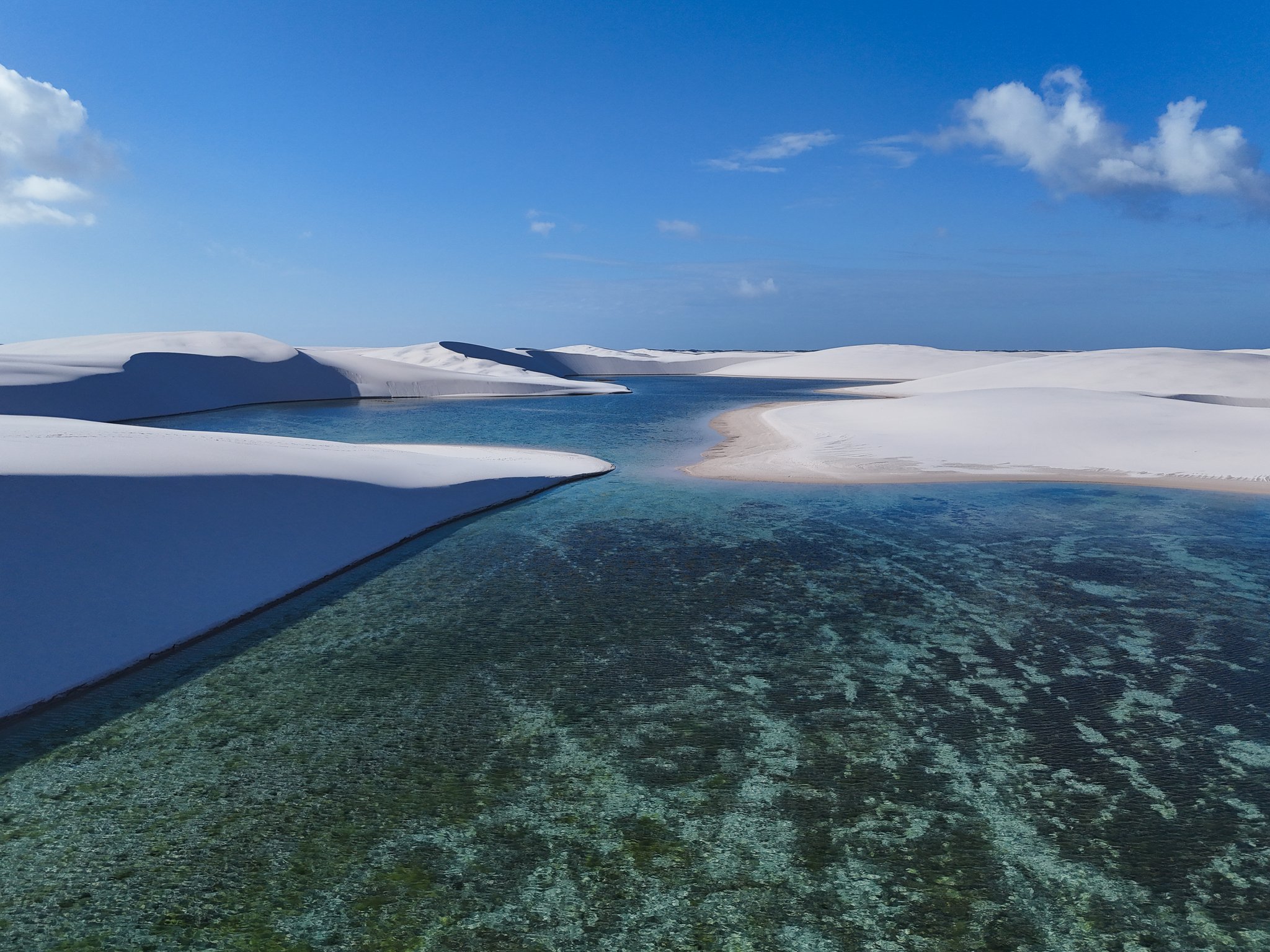 Turquoise water, white sand — sculpted by wind and rain into something otherworldly. Aerial view of a crystal-clear turquoise lagoon nestled between sweeping white sand dunes under a deep blue sky on a Lençóis Maranhenses trip