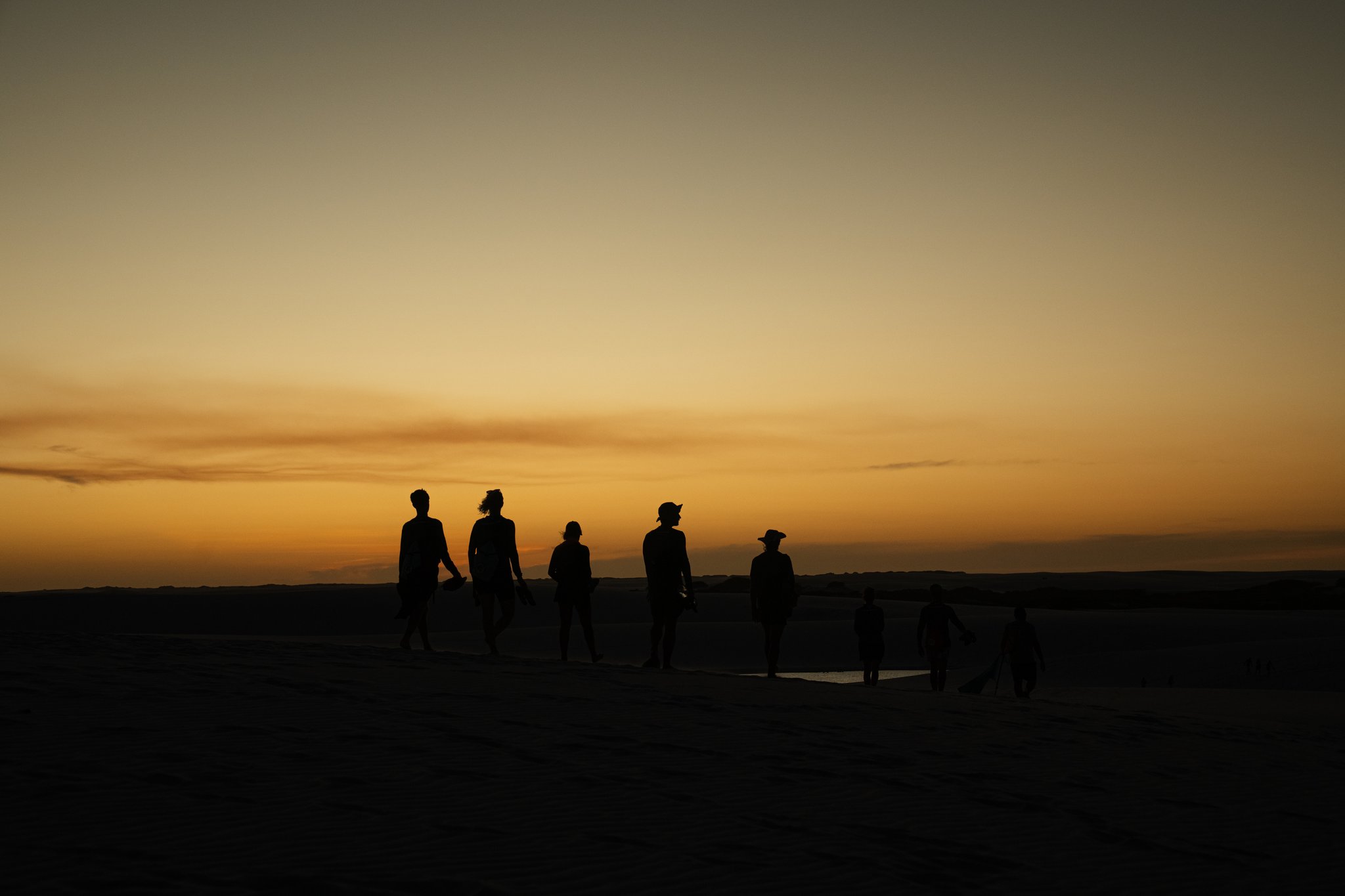 Seven silhouettes, one blazing horizon — the dunes at dusk. Silhouettes of a group of travelers walking across open dunes during a Lençóis Maranhenses trip, set against a vivid amber and orange sunset sky