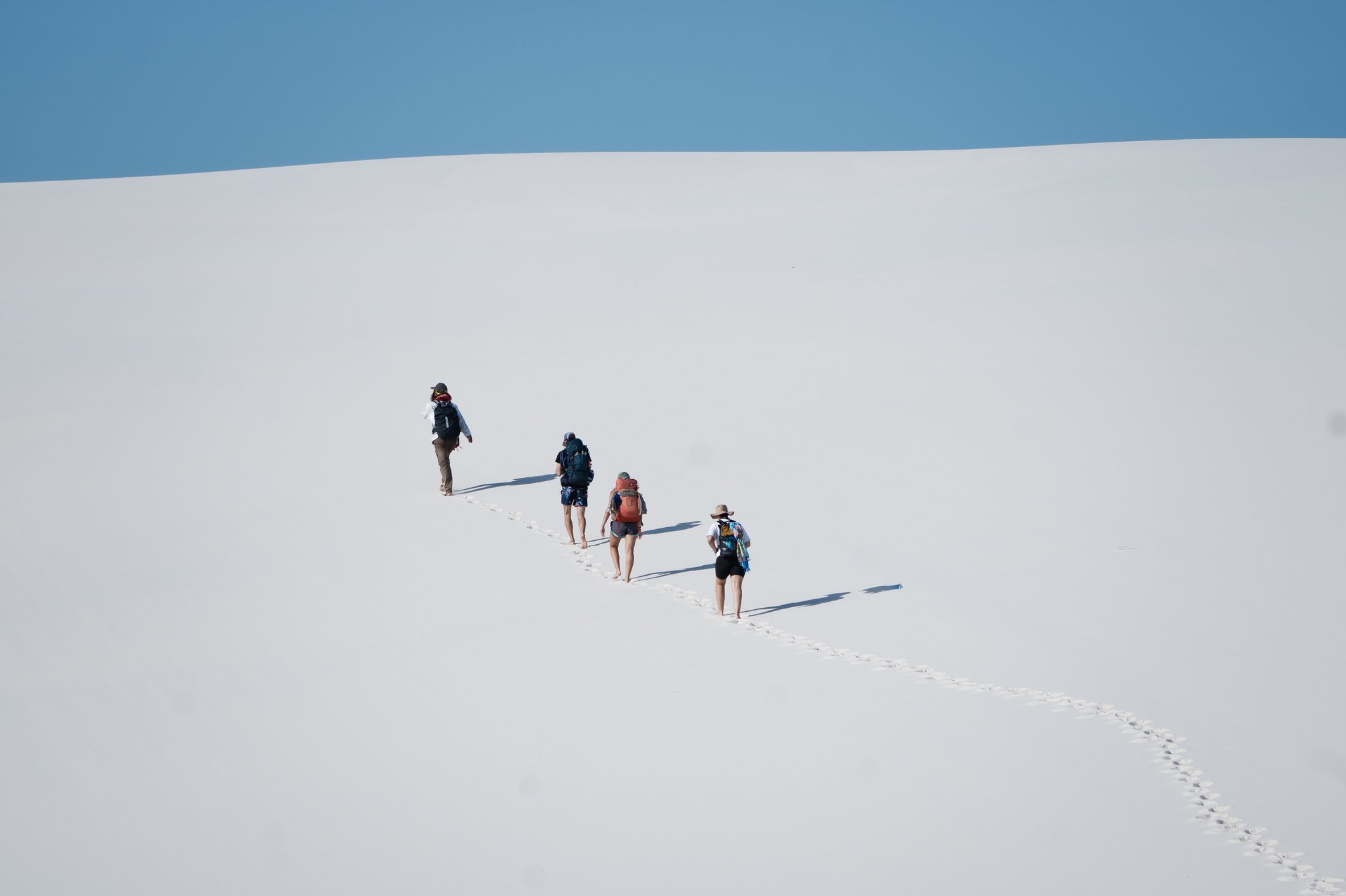 Four figures, one immaculate slope — the dunes reduce everything to its essentials. Four backpackers climbing a vast white sand dune single-file on a Lençóis Maranhenses trip, leaving a trail of barefoot footprints under a clear blue sky