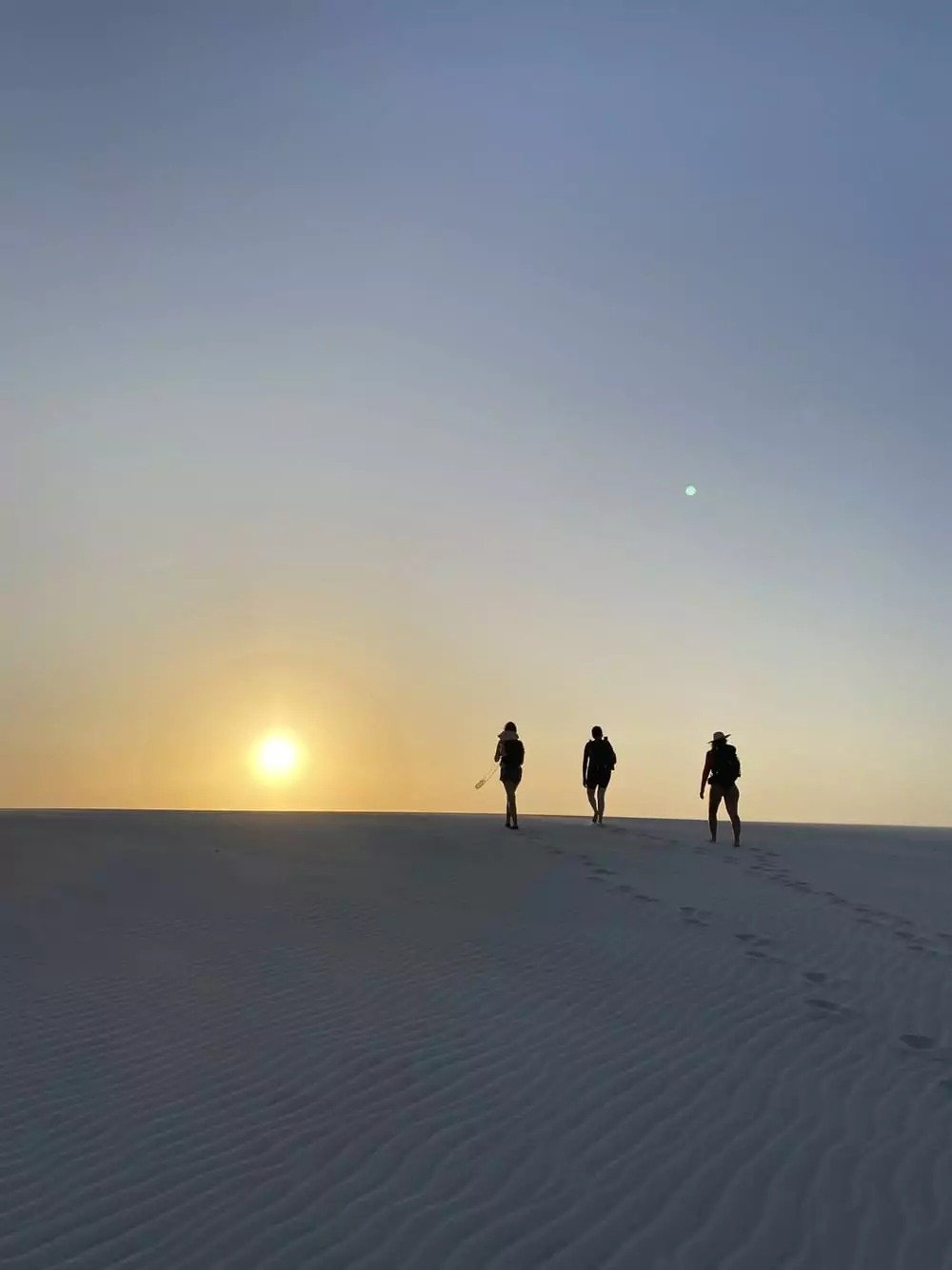 Three silhouettes, one burning sun — walking straight into the horizon. Three travelers silhouetted against a golden sunset sky on a vast white sand dune during a Lençóis Maranhenses trip, footprints trailing behind them