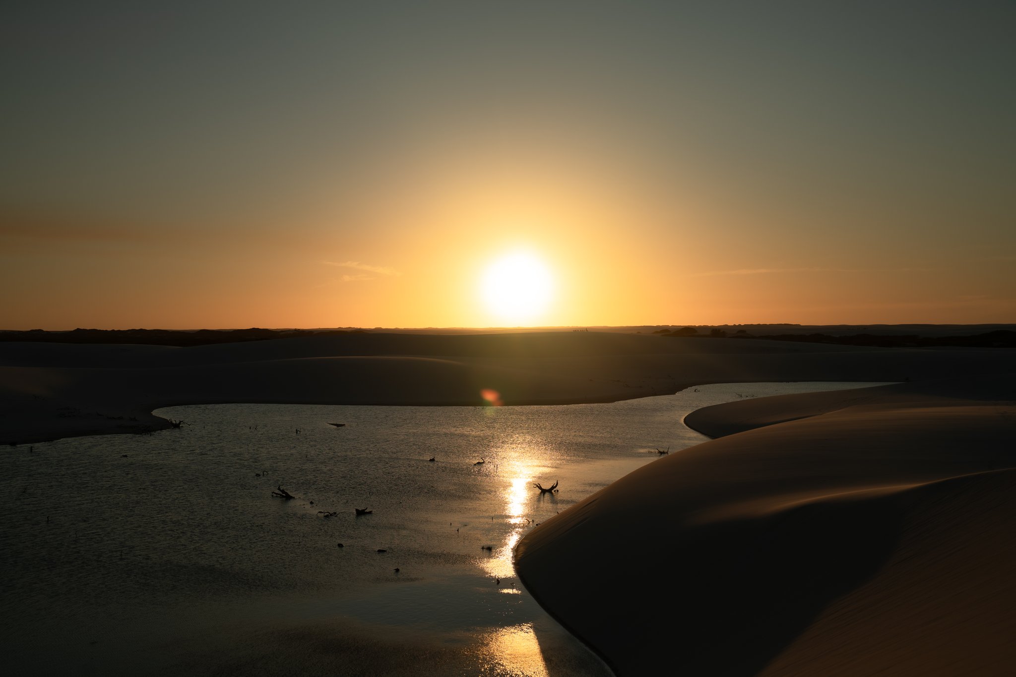 Last light burning across the dunes, the lagoon catching fire. Sun setting on the horizon over a shallow lagoon nestled between dark sand dunes on a Lençóis Maranhenses trip, with birds silhouetted on the glassy water