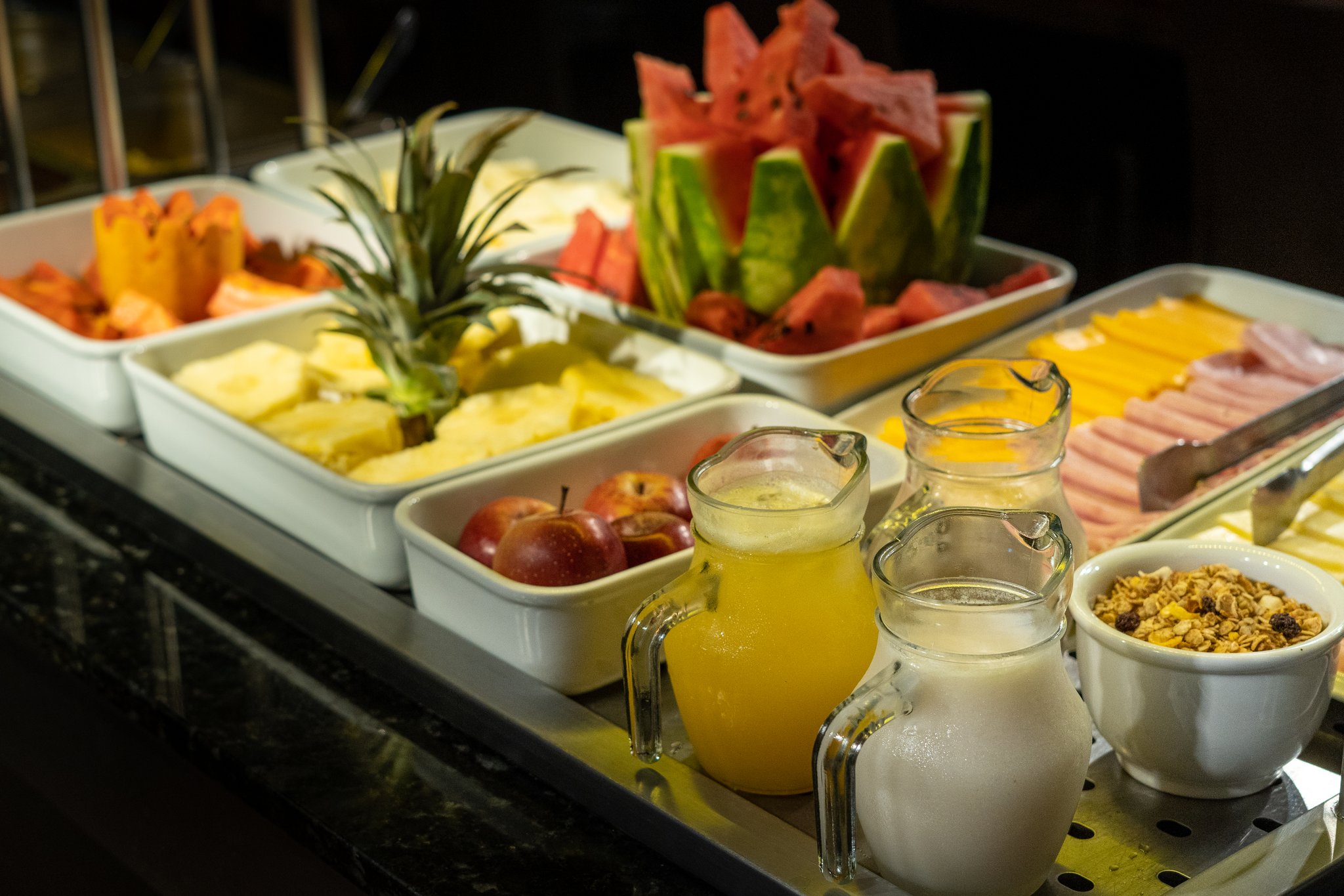 A generous breakfast buffet aboard a luxury Amazon cruise in Brazil, featuring trays of sliced pineapple, watermelon, apples and papaya alongside pitchers of fresh juice, milk, sliced cold cuts, cheese and a bowl of granola